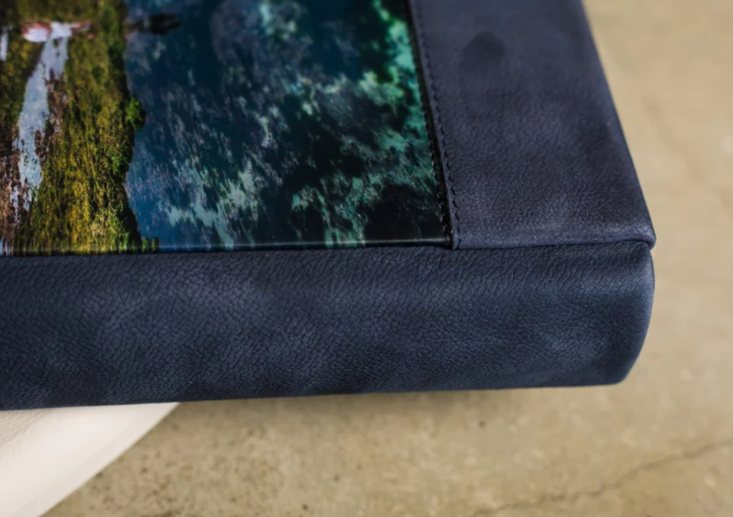 Close-up of a photo book corner with navy blue leather binding and vibrant aerial ocean reef photography print.