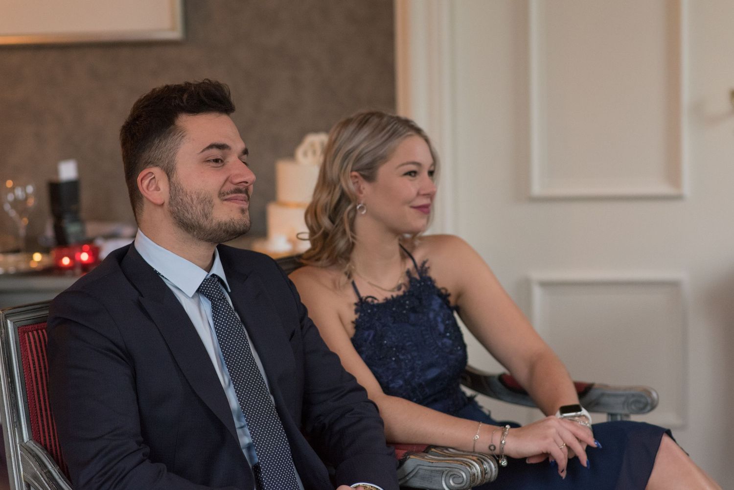 A man and woman in navy formal attire smile while seated at an elegant indoor event.