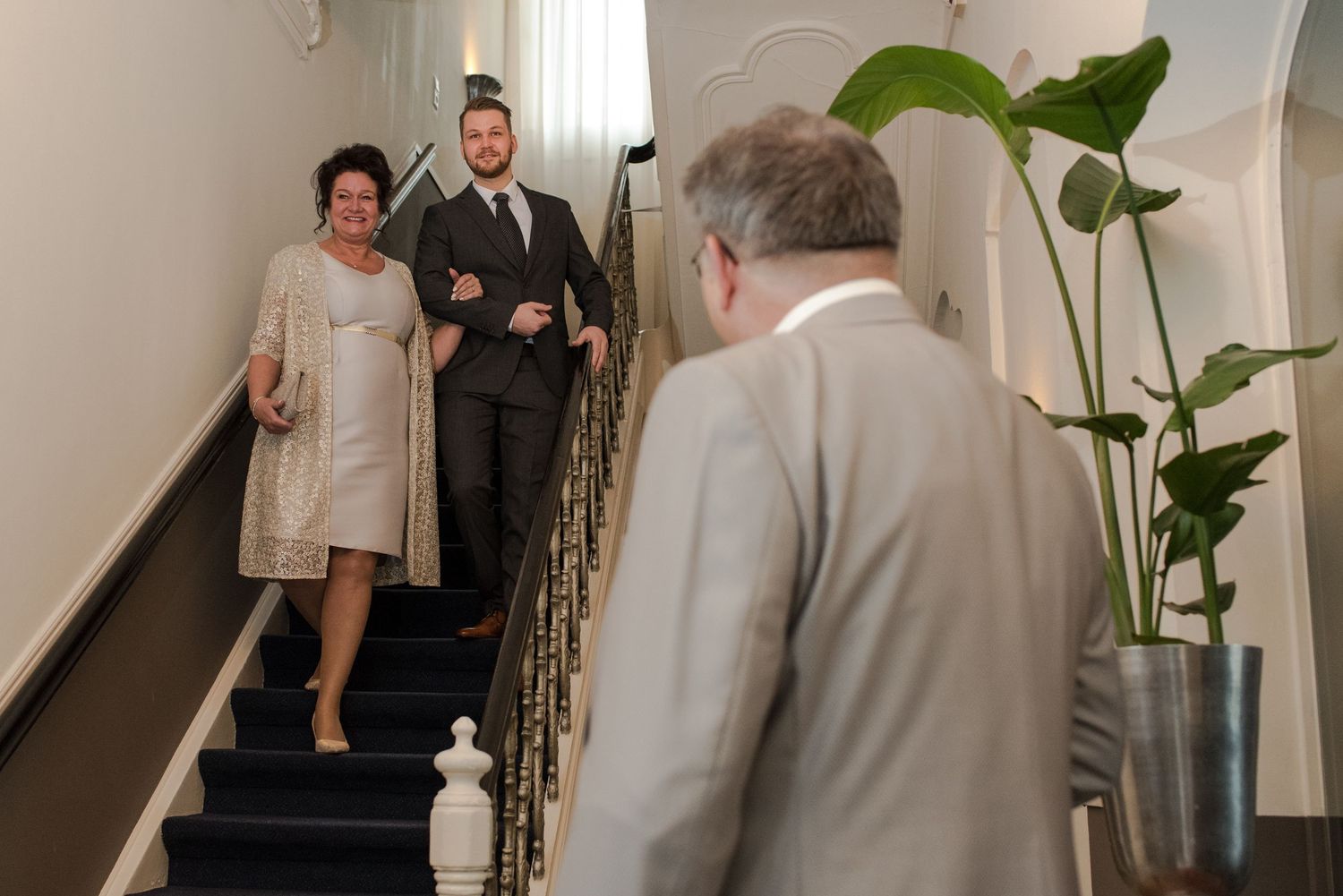 Elegant couple descending a grand staircase at a formal event, greeted by a man in a light gray suit.
