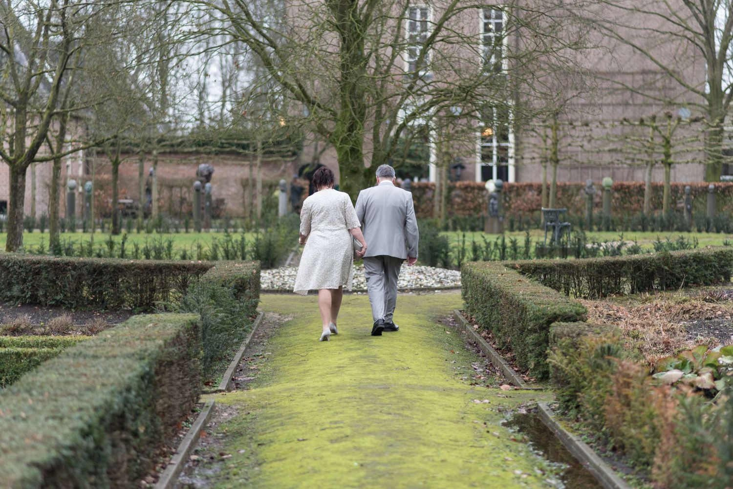 Elderly couple holding hands while walking through a formal garden with manicured hedges and moss-covered path.