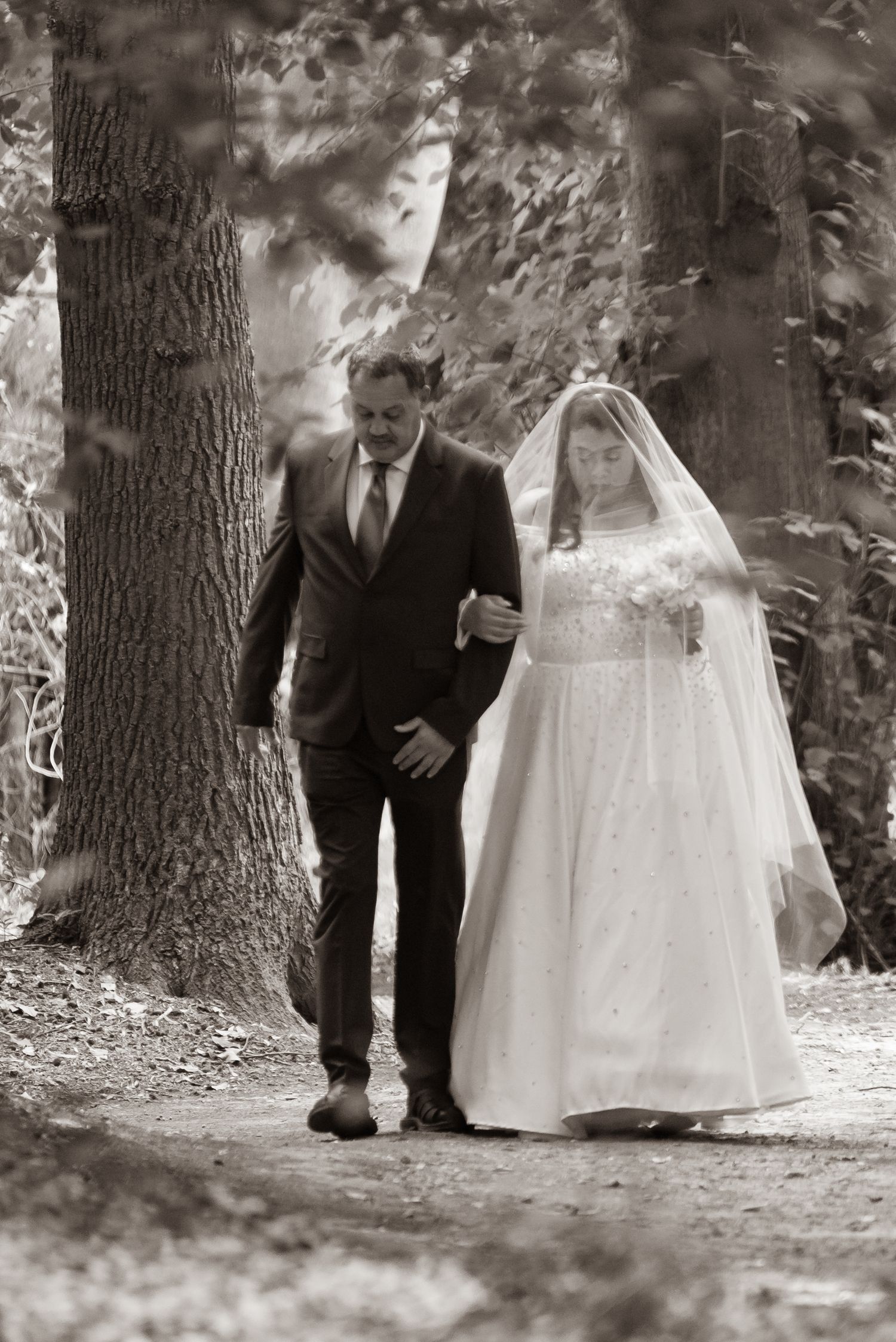 Black and white photo of a bride in white gown and veil walking with a suited man through a wooded forest path.