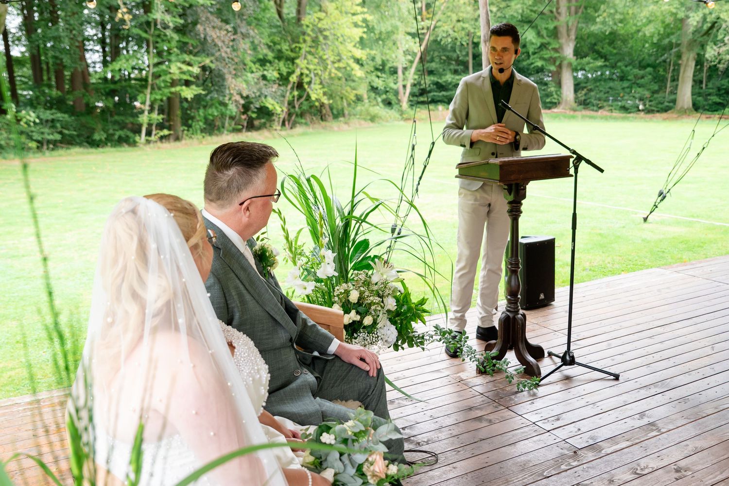 Trouwambtenaar op een houten platform voor  grasveld en bomen bezig met de speech. We zien het bruidspaar dat naar hem kijkt van opzij.