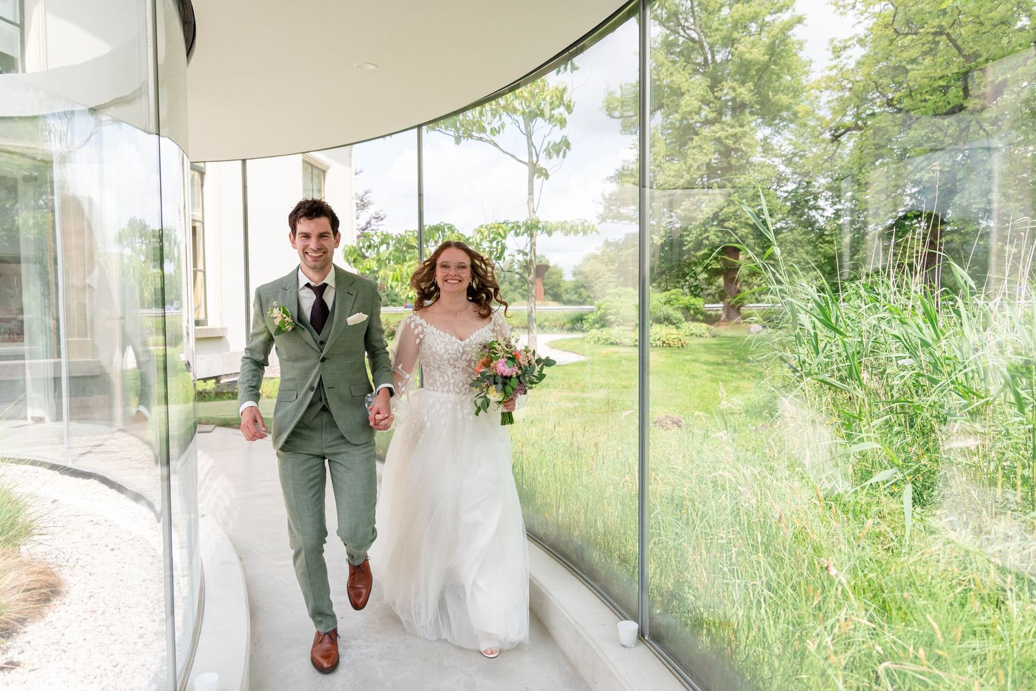 A happy bride and groom in a sage green suit walk hand-in-hand through a modern glass corridor.