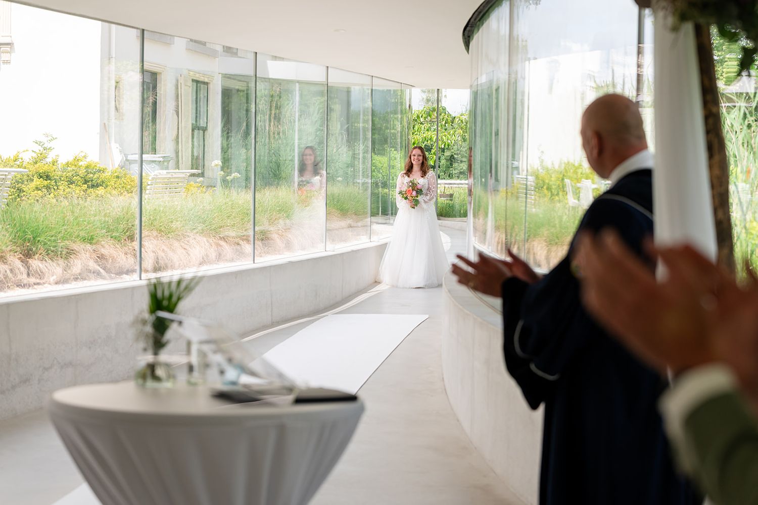 Bride in white gown holding bouquet walks down aisle as guests applaud in elegant glass-walled wedding venue.