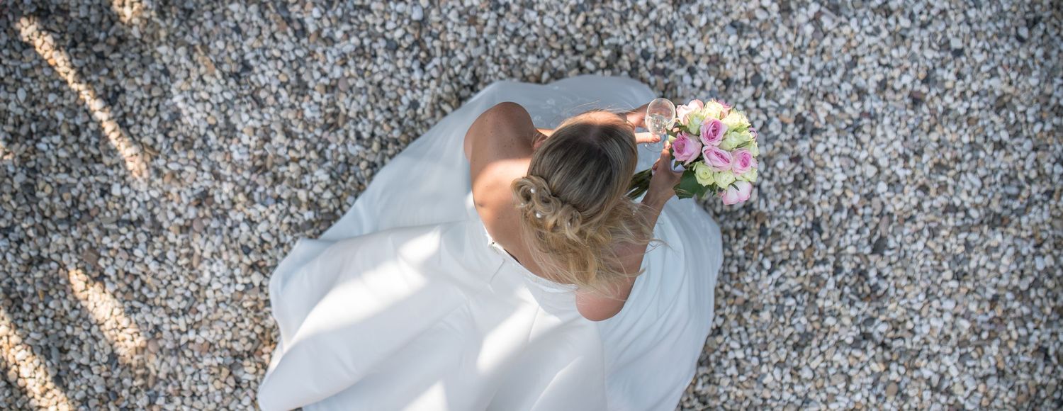 Aerial view of a bride in a white dress with floral hair accessory standing on a pebble beach.