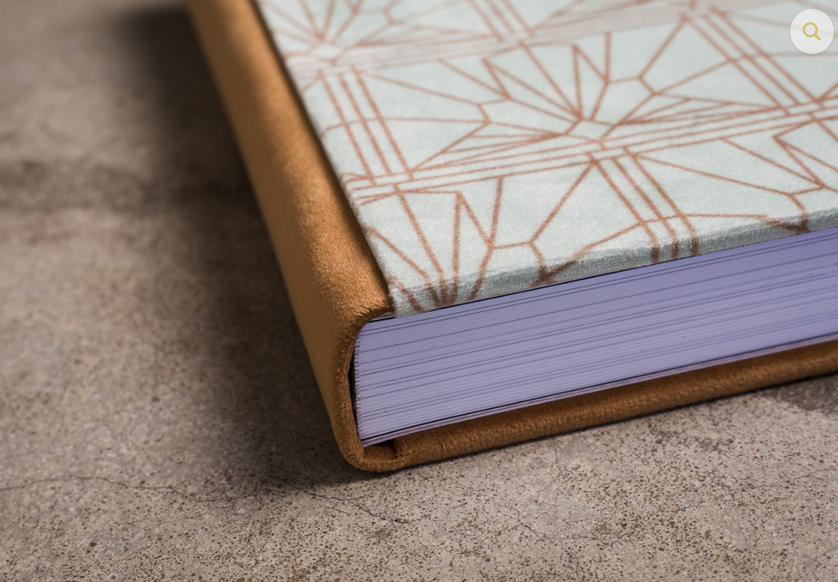 Close-up of a hardcover book with tan suede spine, geometric mint and copper patterned cover, and lavender page edges on a gray surface.