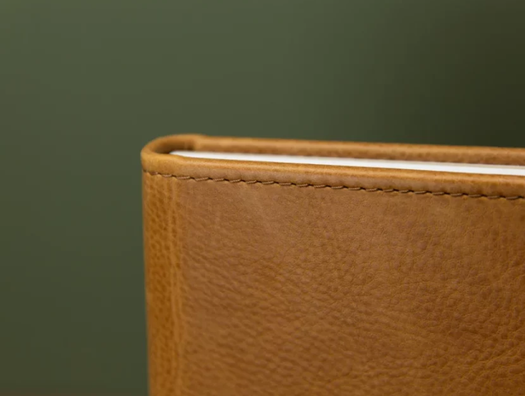 Close-up of a tan leather wallet corner showing fine stitching and pebbled texture against a dark green background.