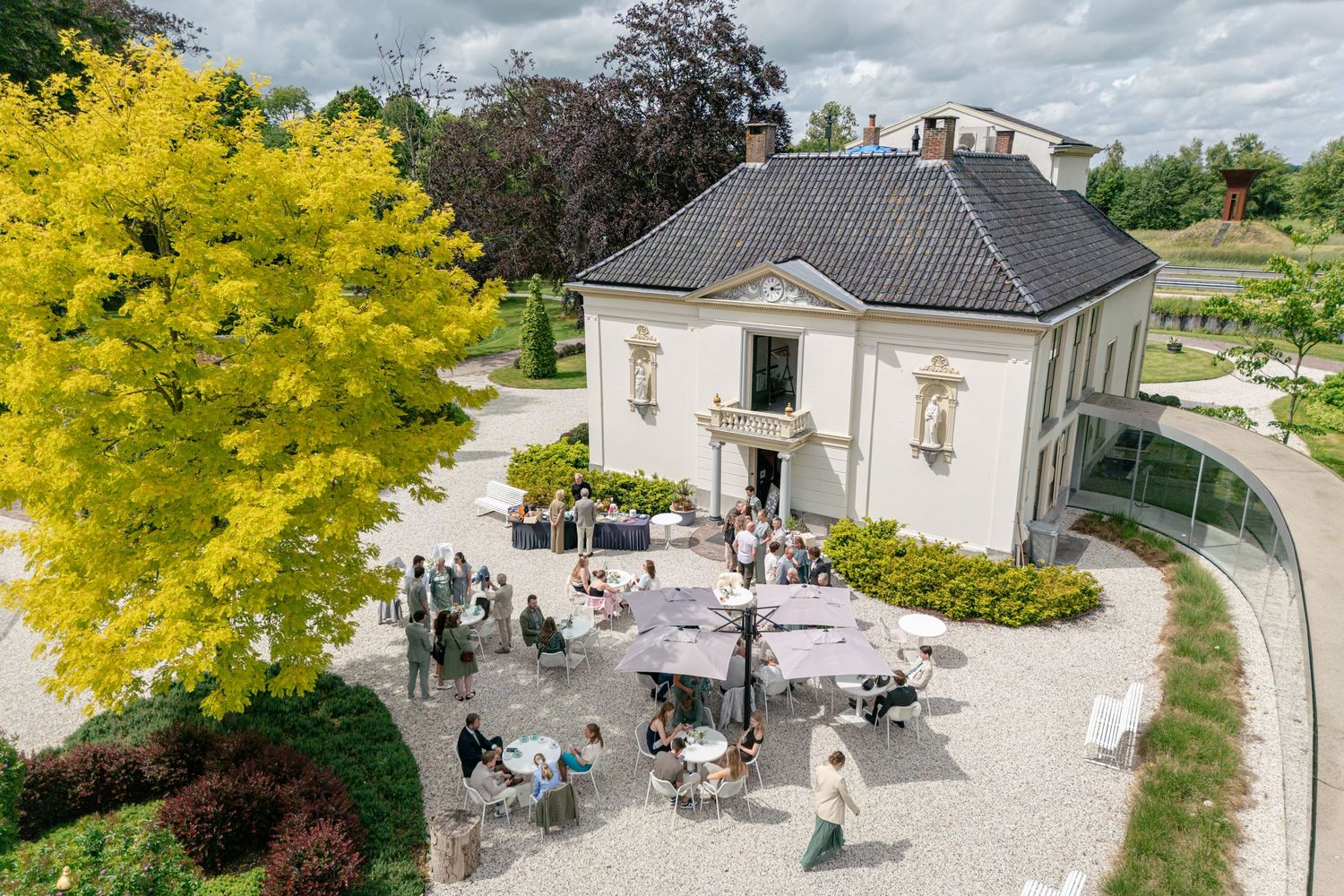 Aerial view of elegant villa with outdoor cafe seating, surrounded by lush green trees and manicured gardens.