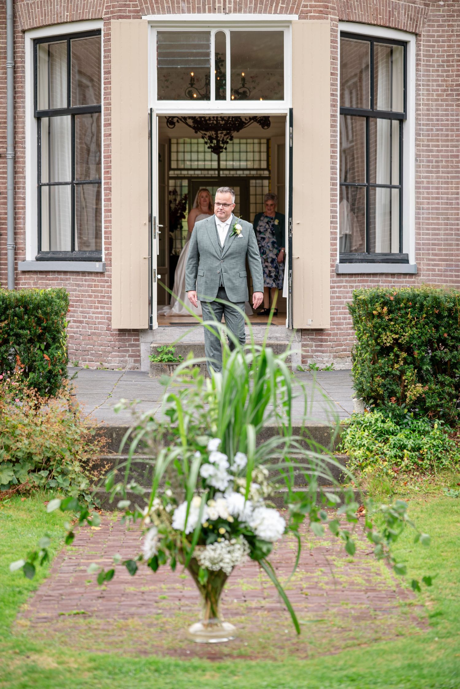 De bruidegom loopt vanuit Klein Soestdijk de tuin in waar de trouwceremonie plaatsvindt. Op de voorgrond een bloemstuk dat in de tuin staat.