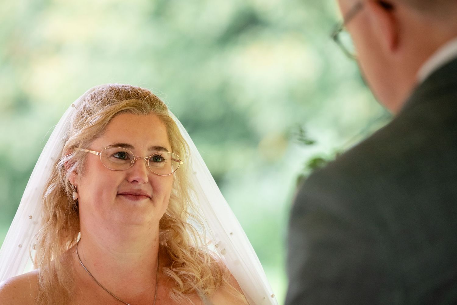 Een leifdevolle blik van een bruid naar haar bruidegom tegen een wazige achtergrond van groene bomen.