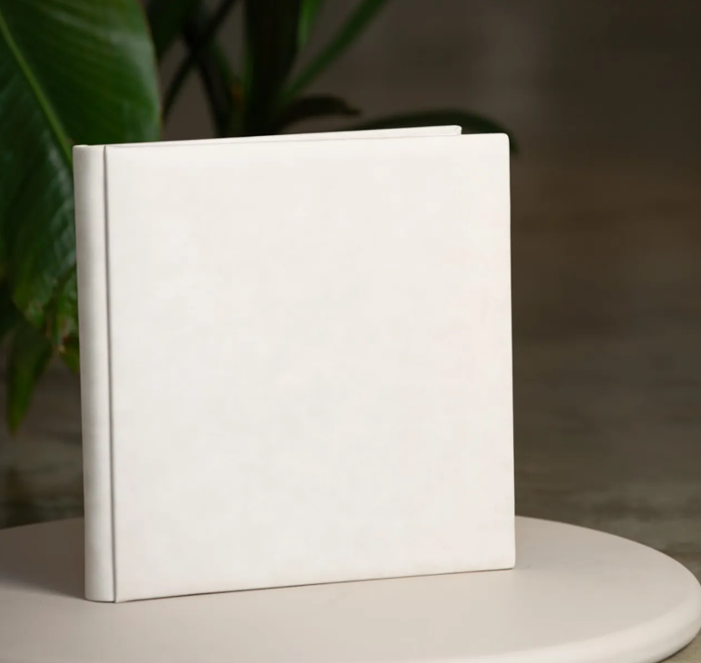 A white square photo album with a clean minimalist design, displayed on a white table with green plants in the background.
