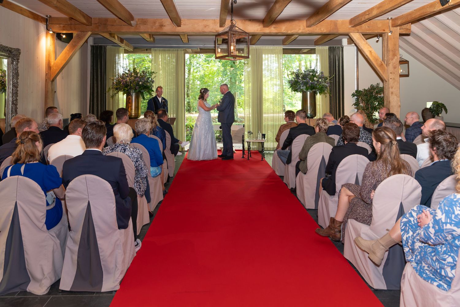 Indoor wedding ceremony with guests seated on white chairs along a red carpet aisle, couple at altar in a rustic venue.