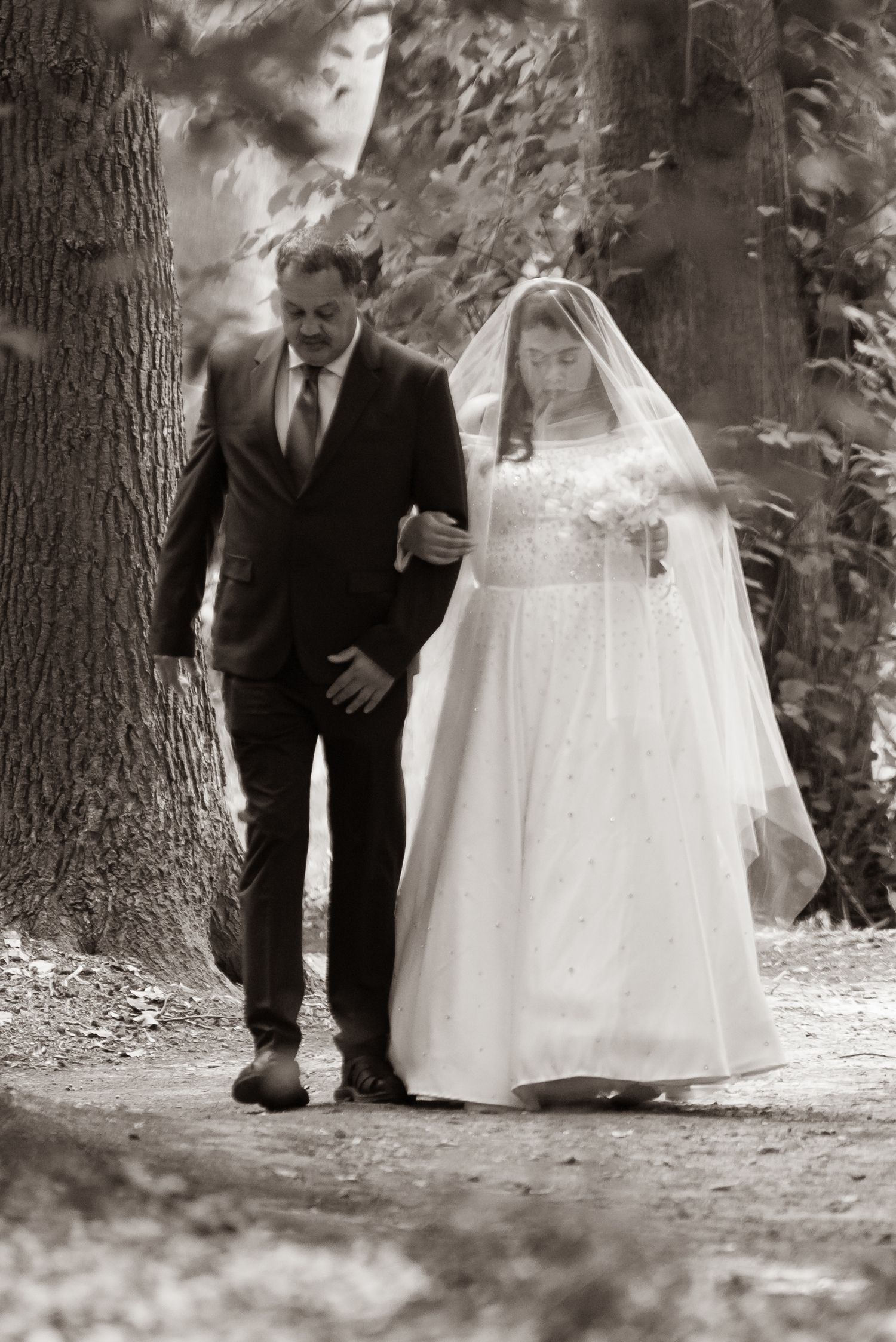 A bride in a white gown and veil walks arm-in-arm with a suited man through a wooded path, sepia tone.
