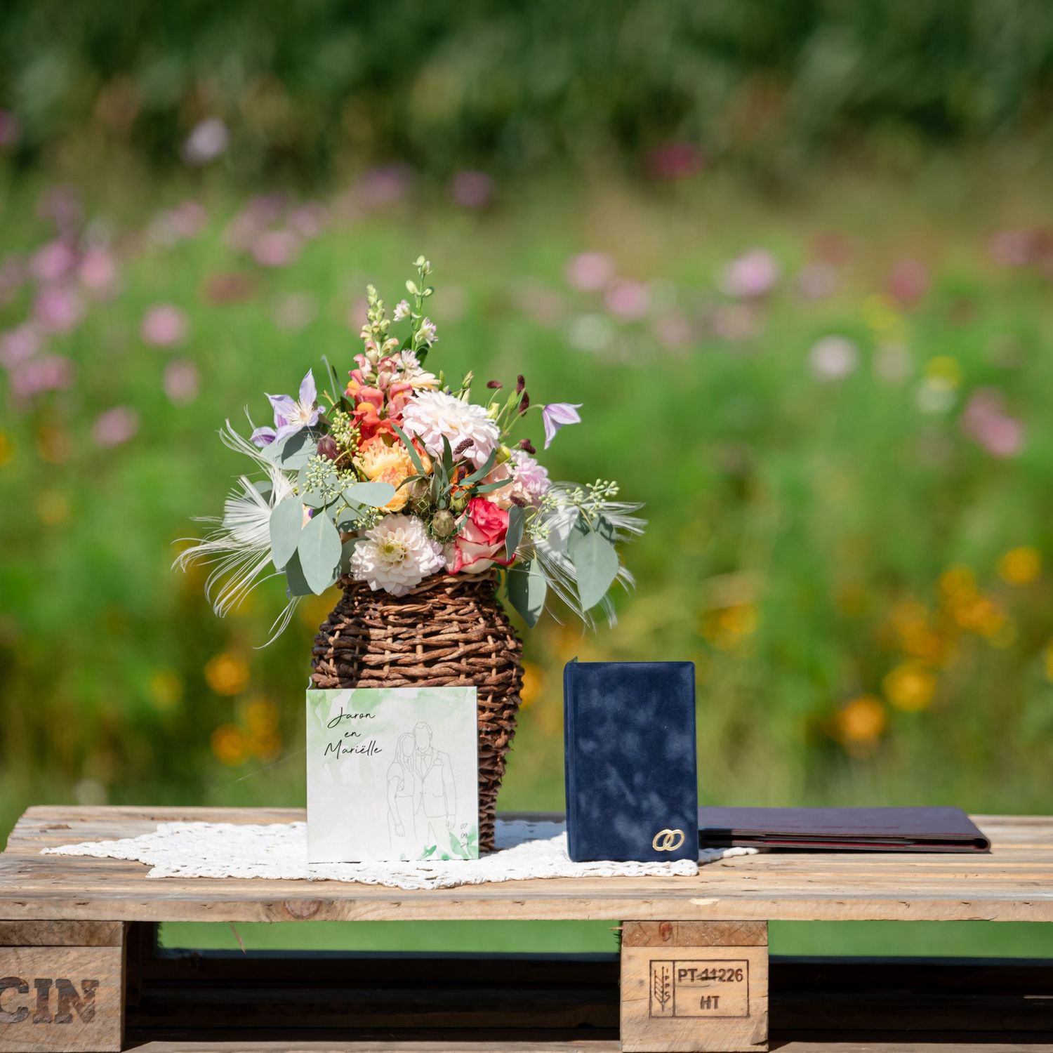 Wicker basket of pastel wildflowers on a rustic pallet table beside a navy ring box and wedding card, with a blurred meadow behind.