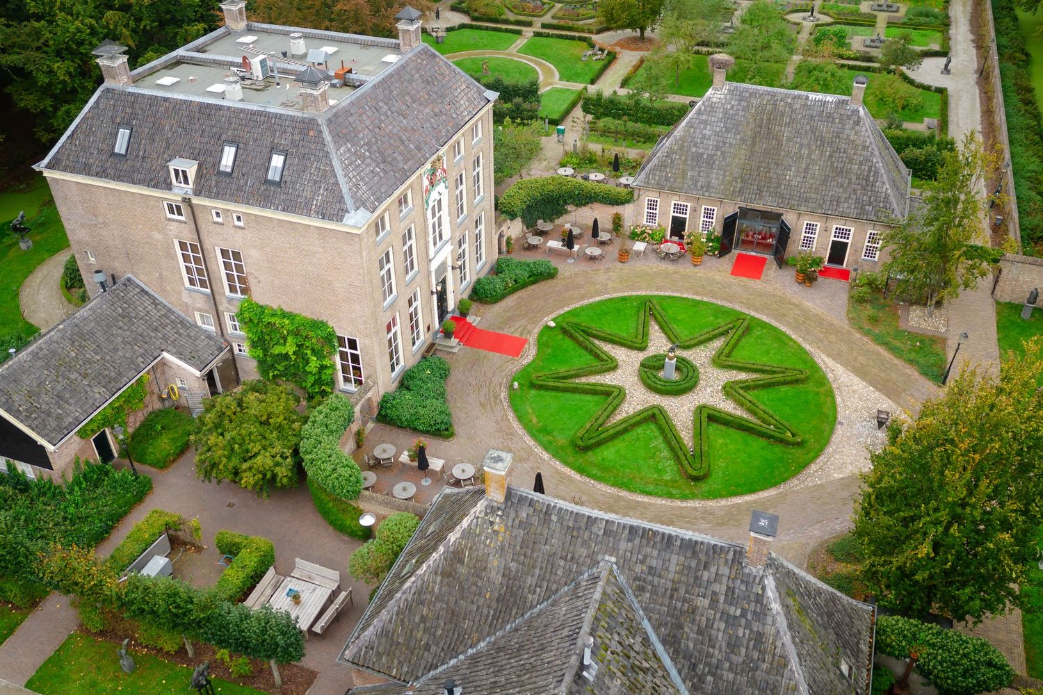 Aerial view of a historic Dutch manor house with ornate star-shaped garden, red carpet entrance, and manicured grounds.