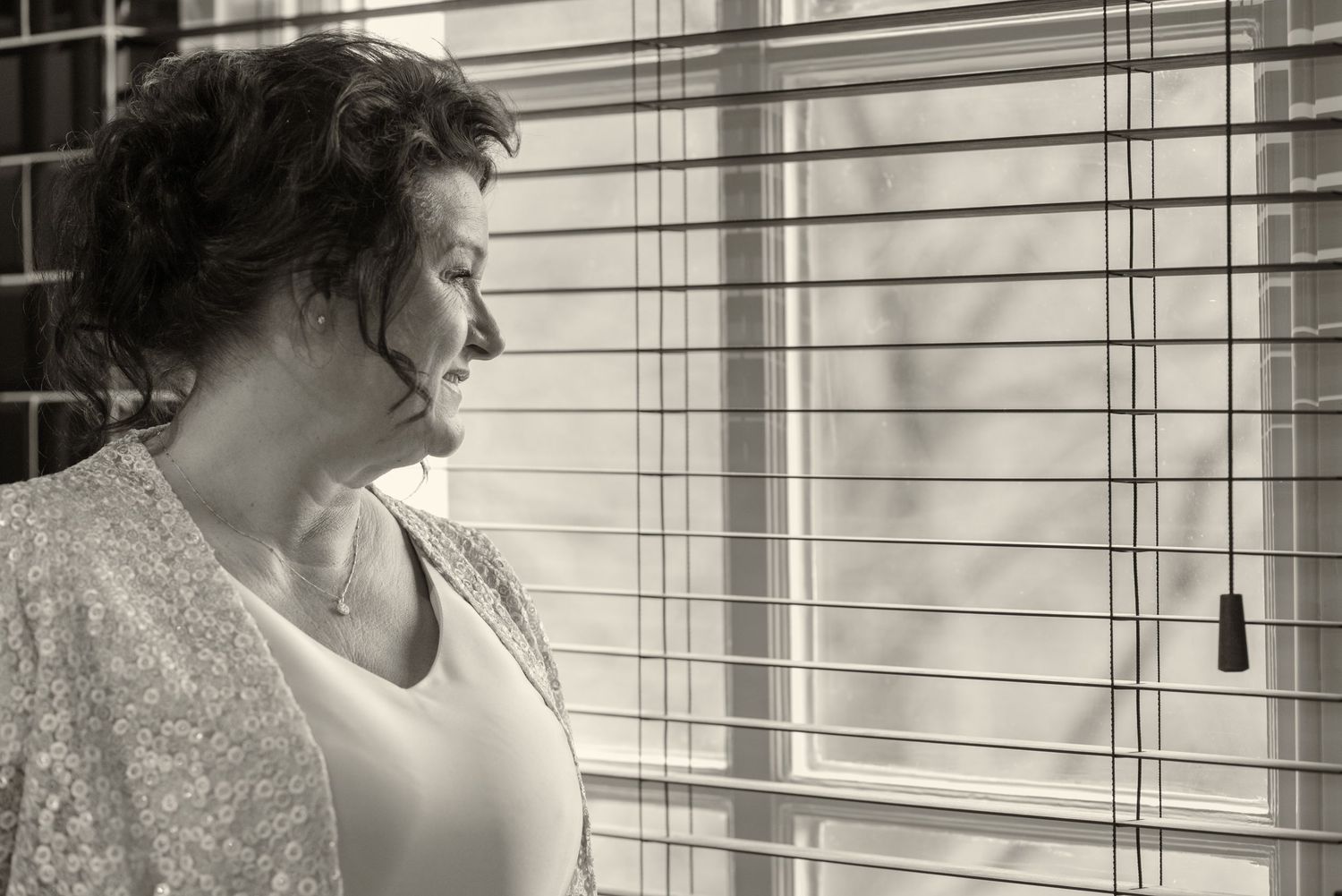 Black and white photo of a woman gazing thoughtfully through window blinds, soft light illuminating her profile.