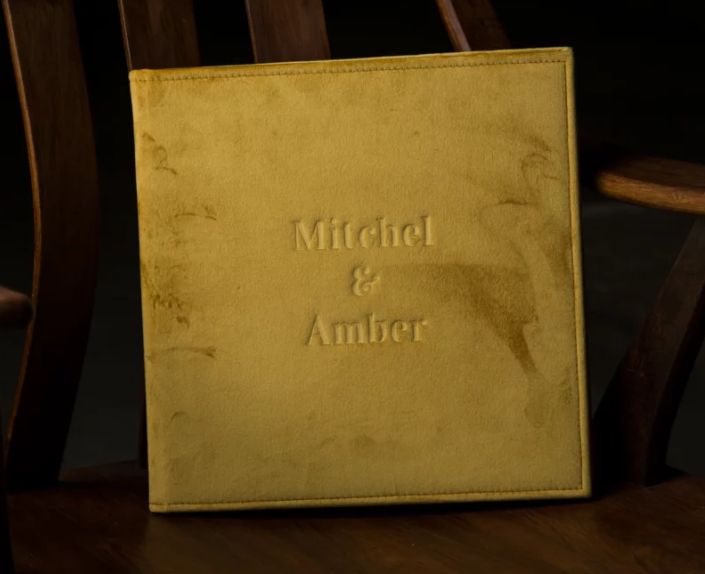Gold embossed wedding photo album with names Mitchel and Amber, leaning against a wooden chair.
