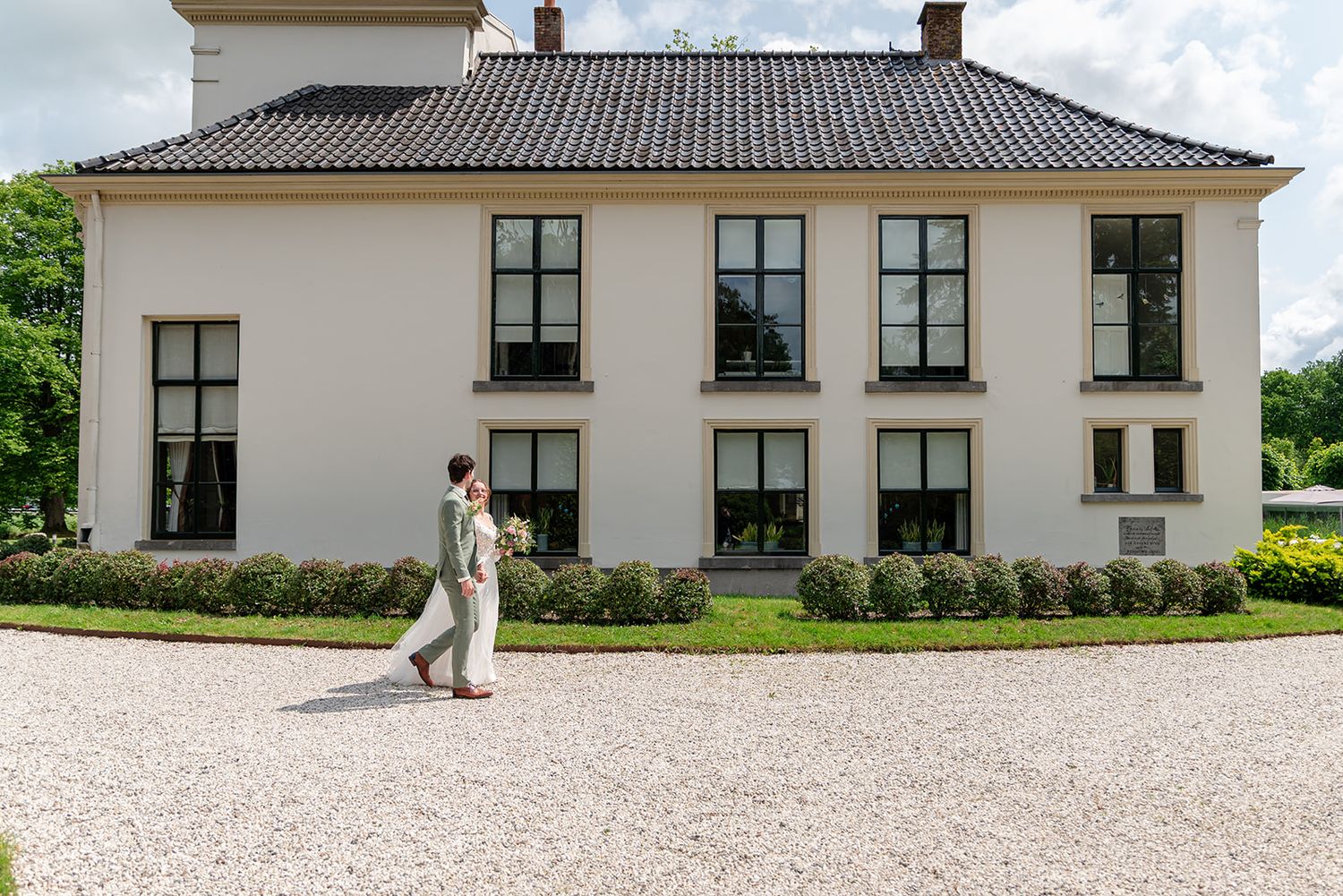 A person walks along a gravel path beside a elegant white Dutch manor house with black-framed windows.