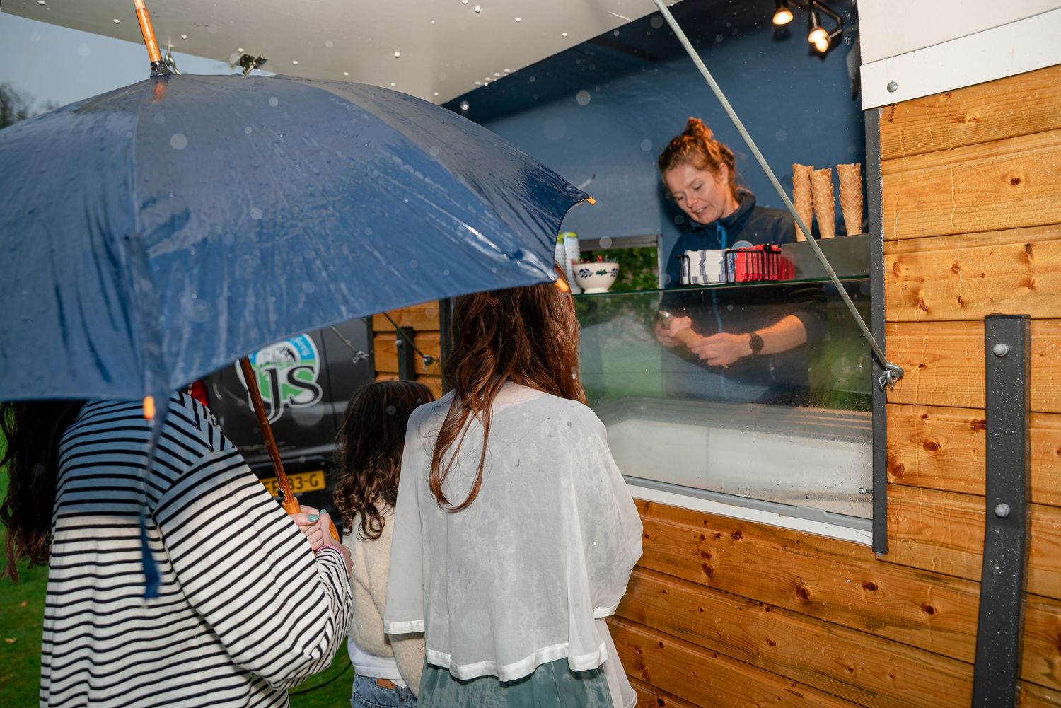 Een ijscowagen in de regen.  Dame in de ijskar schept bolletjes in een hoorntje voor feestelijk geklede wachtende klanten met een paraplu.