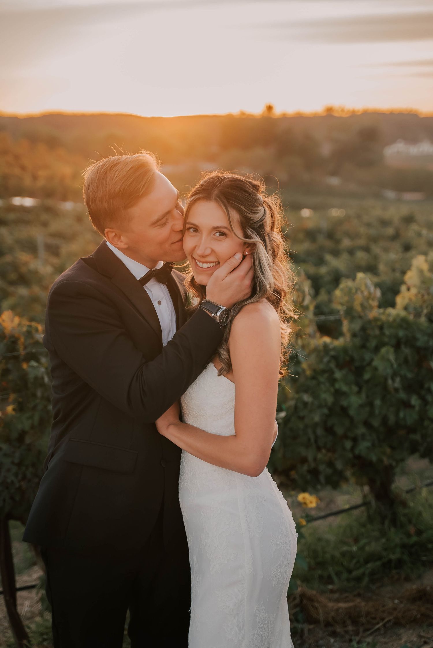 Couple embracing at golden hour sunset in vineyard, groom in tuxedo kissing bride in white gown on cheek.