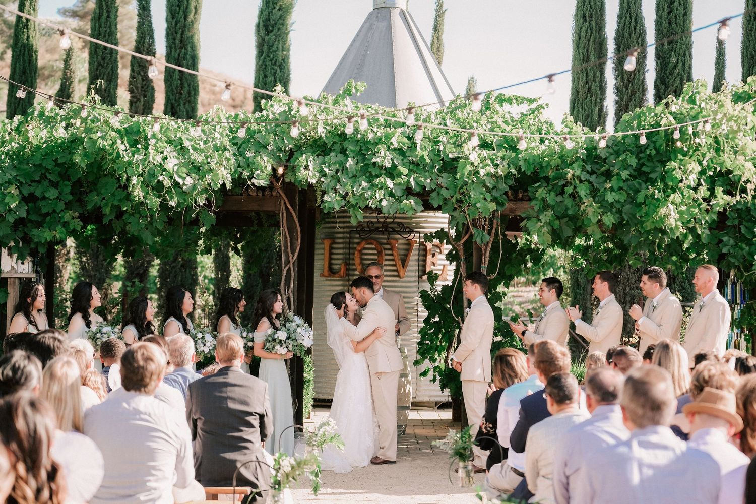 Couple kissing at altar during outdoor wedding ceremony under green vine pergola with guests seated in rows.