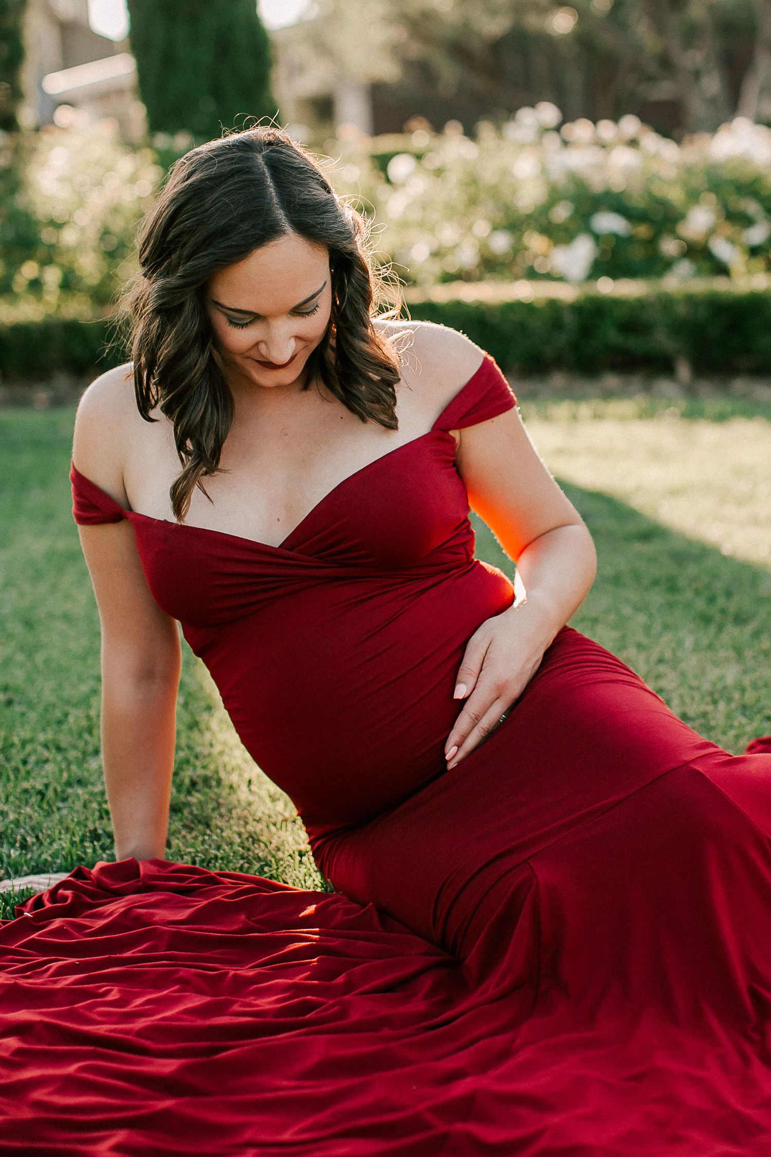 Pregnant woman in elegant red off-shoulder gown sitting on grass, cradling baby bump during outdoor maternity photoshoot.