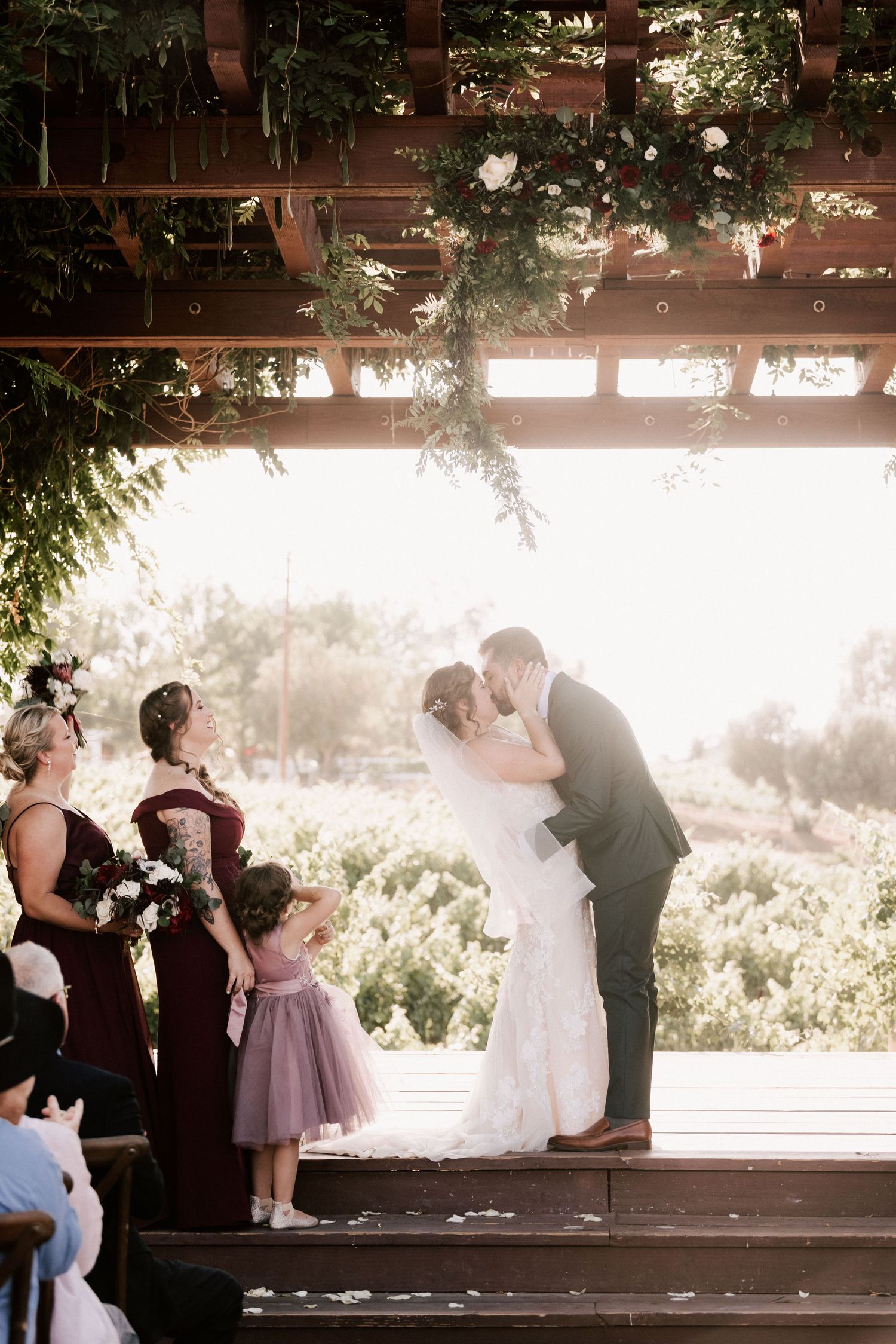 Bride and groom share first kiss at outdoor wedding ceremony under floral pergola with bridesmaids in burgundy dresses.