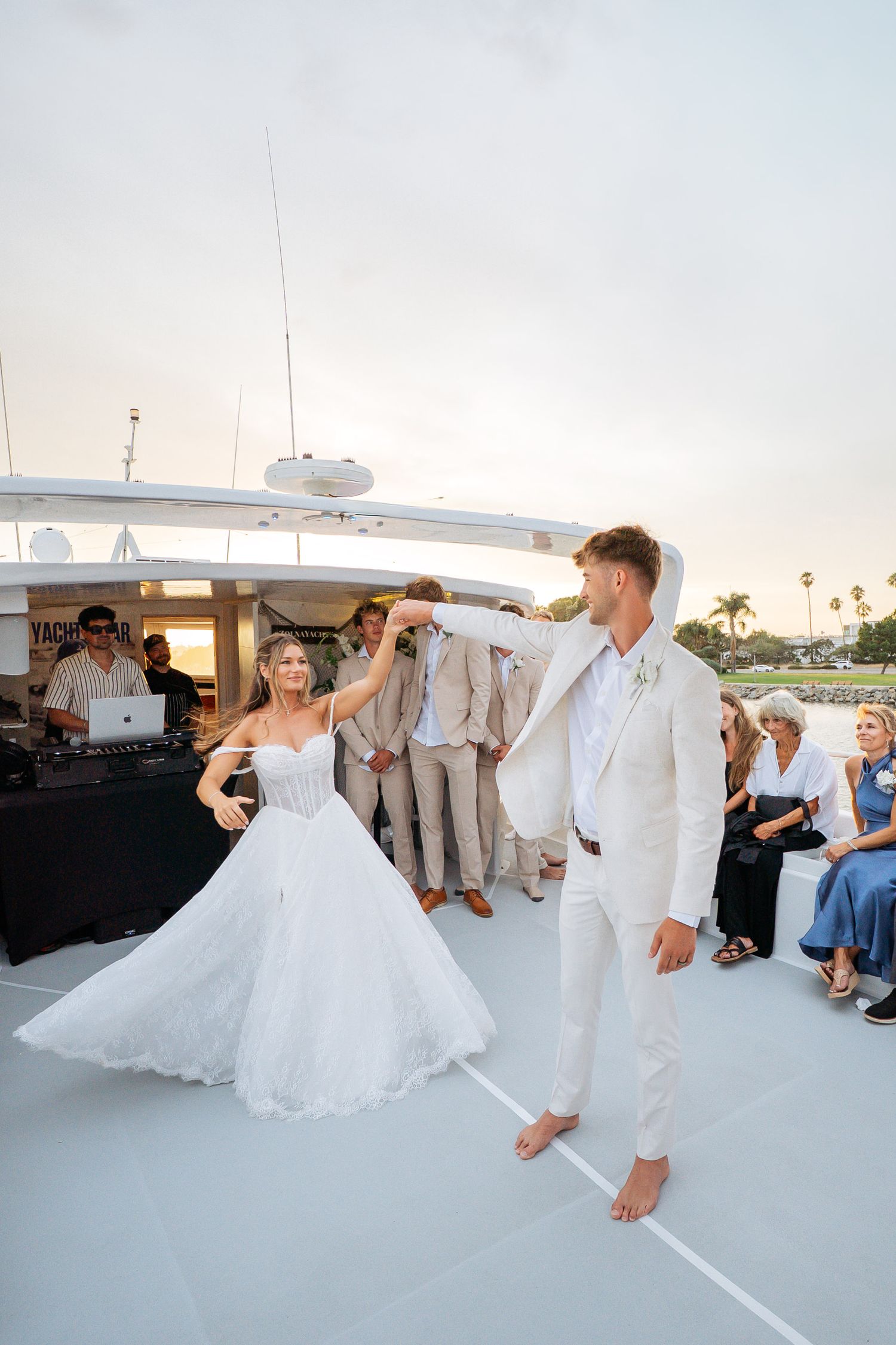 Bride and groom share first dance on yacht deck at sunset wedding reception with guests watching.