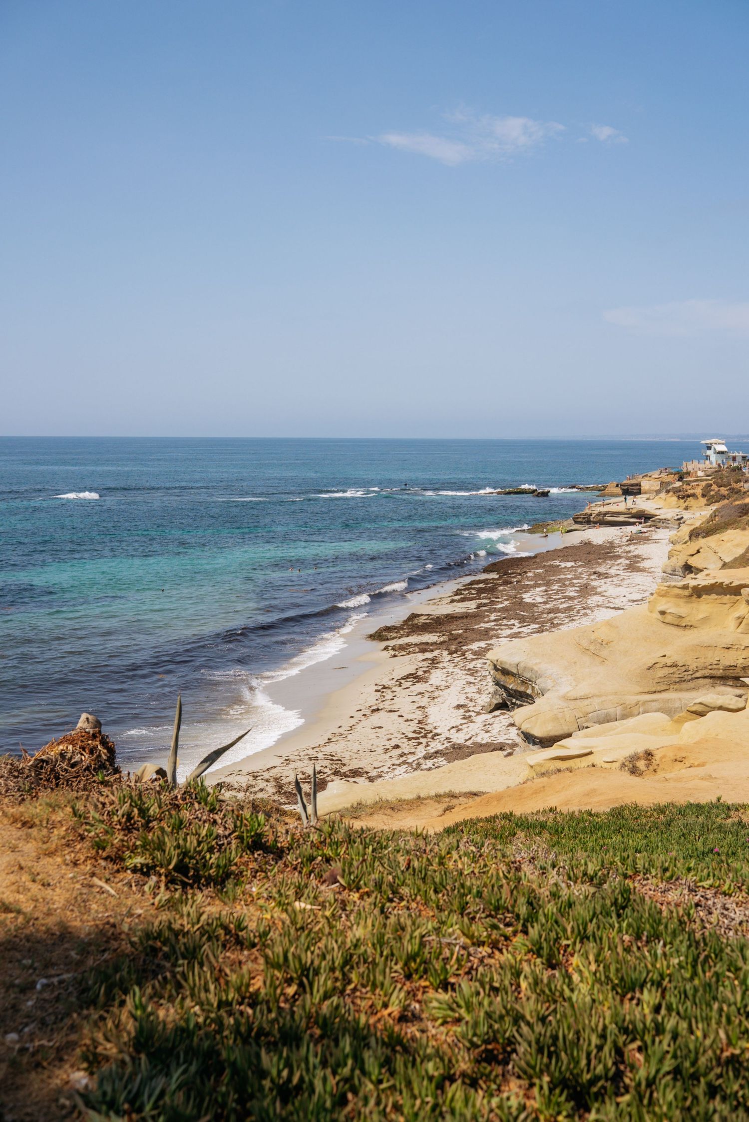 Rocky coastal cliffside overlooking a sandy beach with turquoise ocean waves under a clear blue sky.
