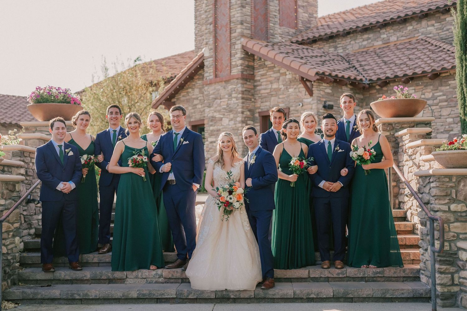 Wedding party in green dresses and navy suits posing on stone steps outside a rustic venue.