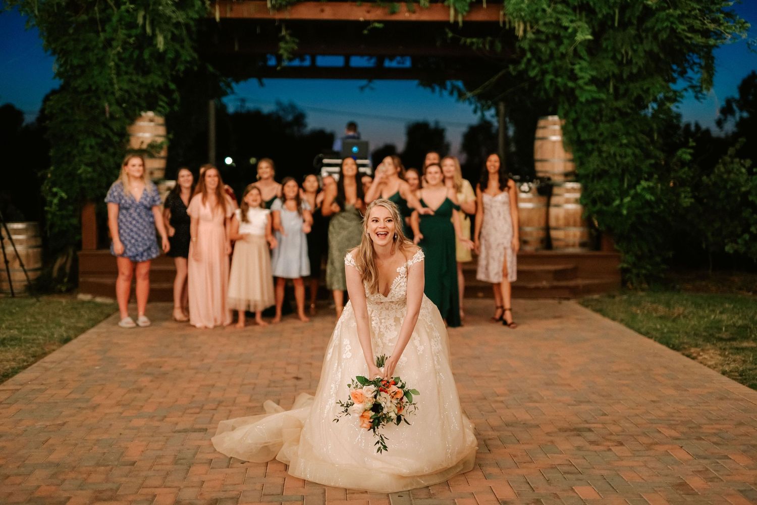 Bride tosses bouquet to bridesmaids in blush and green gowns at outdoor wedding venue with barrel arch backdrop.