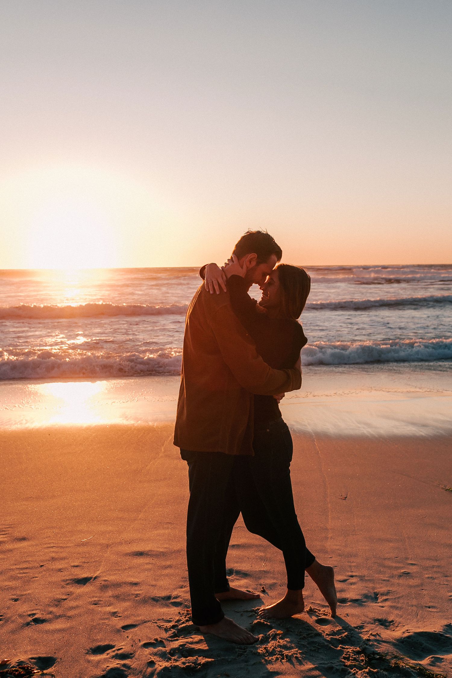Couple silhouetted against a vibrant ocean sunset, embracing barefoot on wet sand.