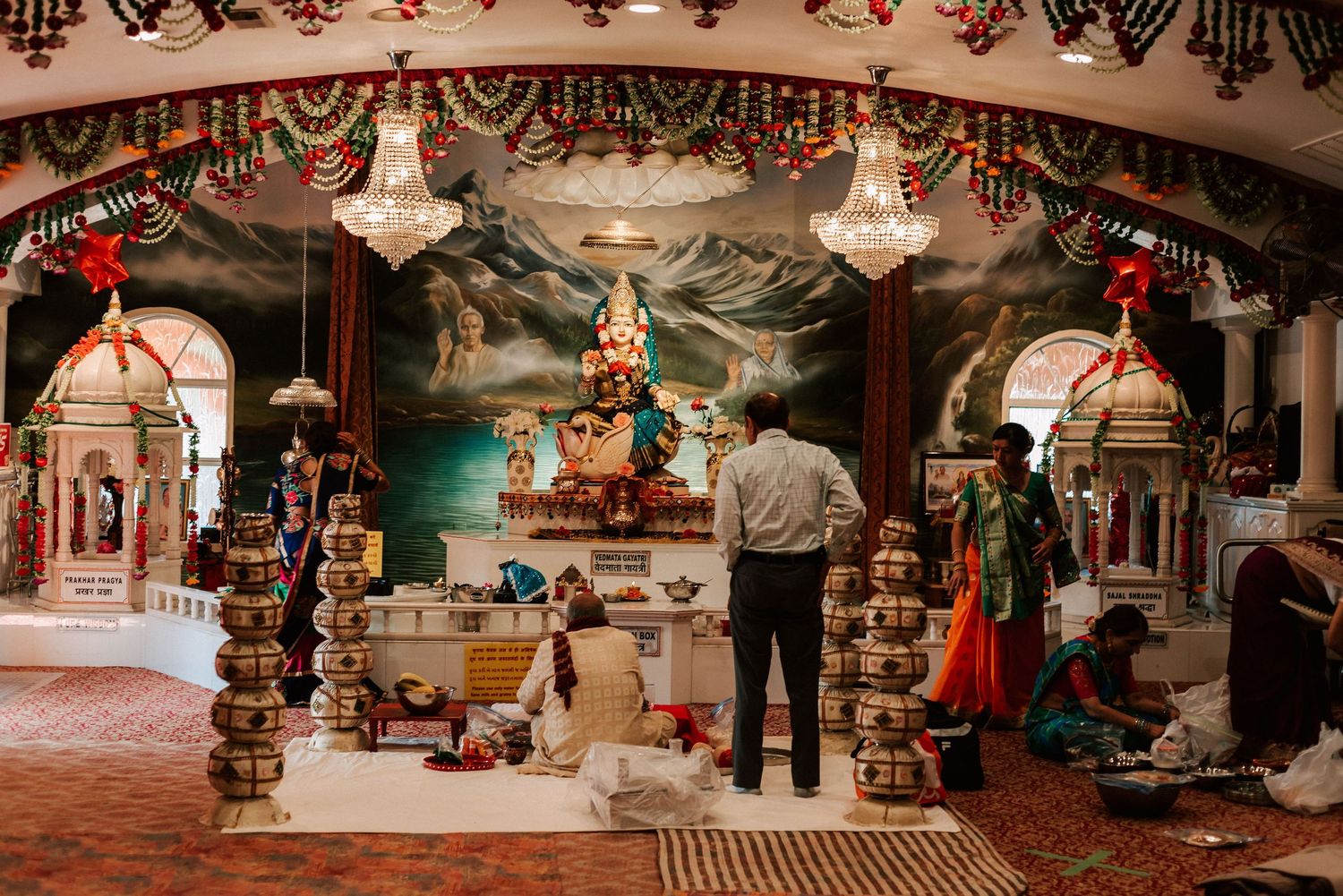 Elaborately decorated Hindu temple interior with chandeliers, floral garlands, and devotees performing puja rituals.