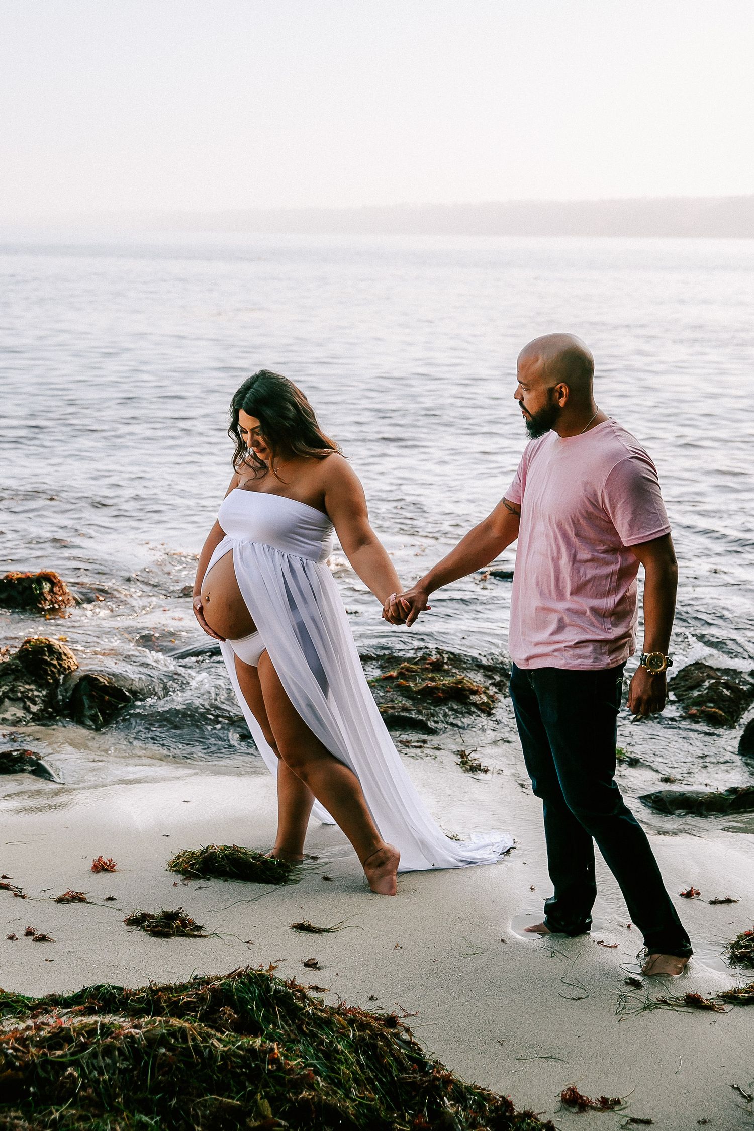 Couple holding hands walking along rocky shoreline, pregnant woman in flowing white maternity dress.