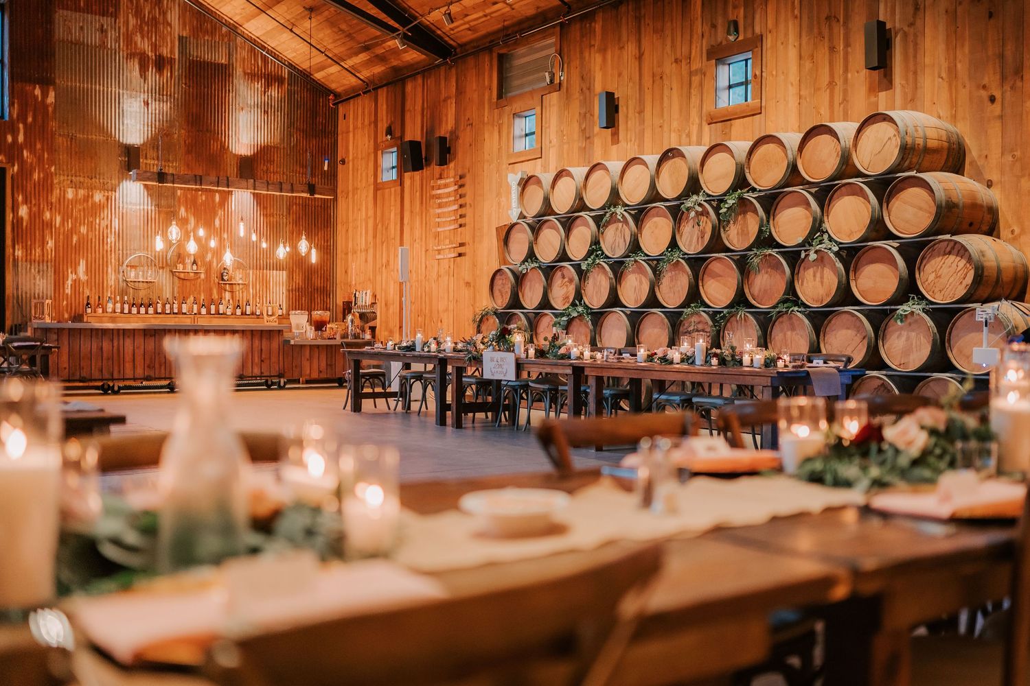 Rustic winery event venue with wooden barrels wall, candlelit dining tables, and warm string lights for elegant wedding reception.