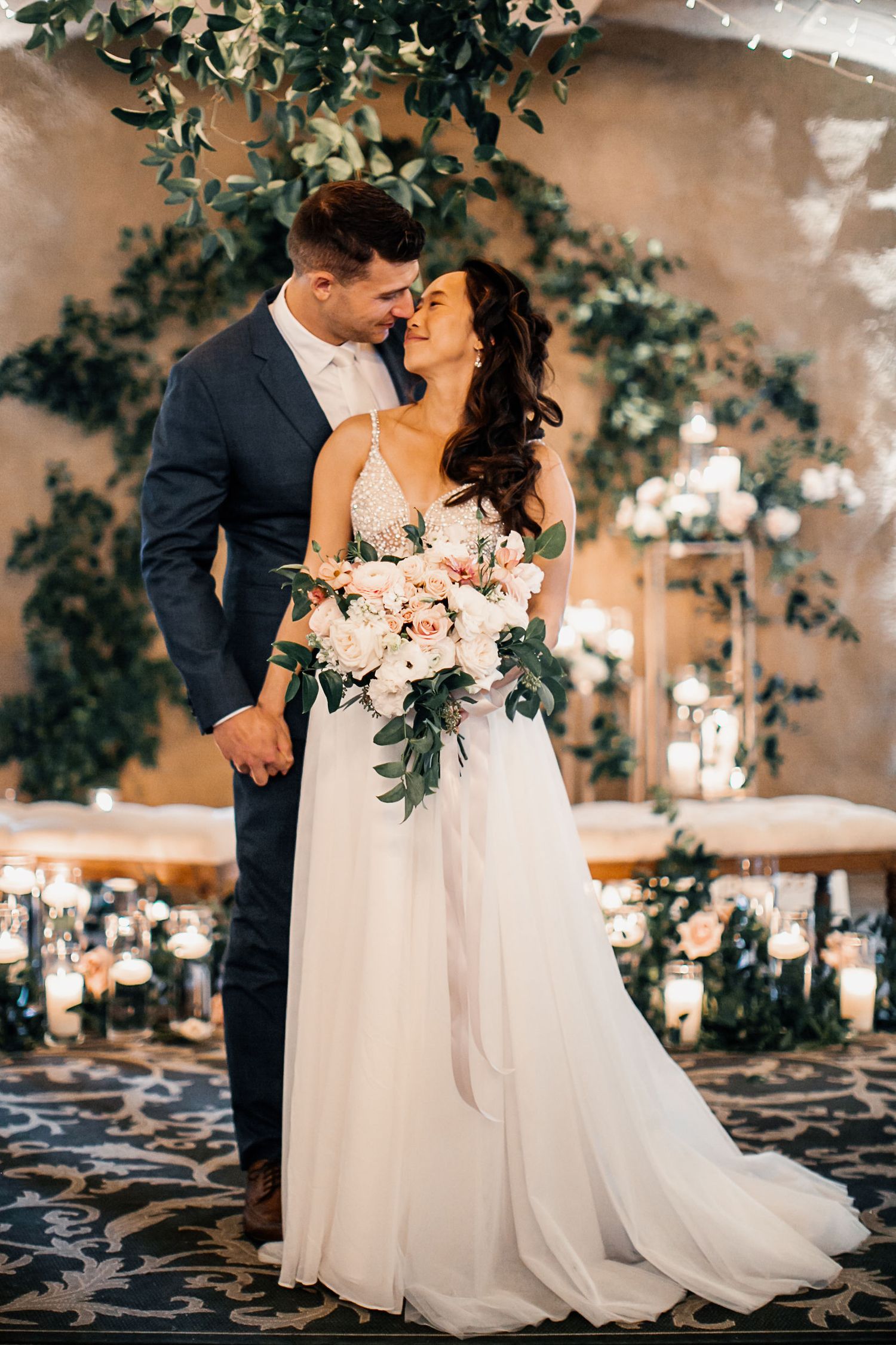 Bride and groom share romantic moment at elegant wedding venue with floral greenery and candlelight decor.