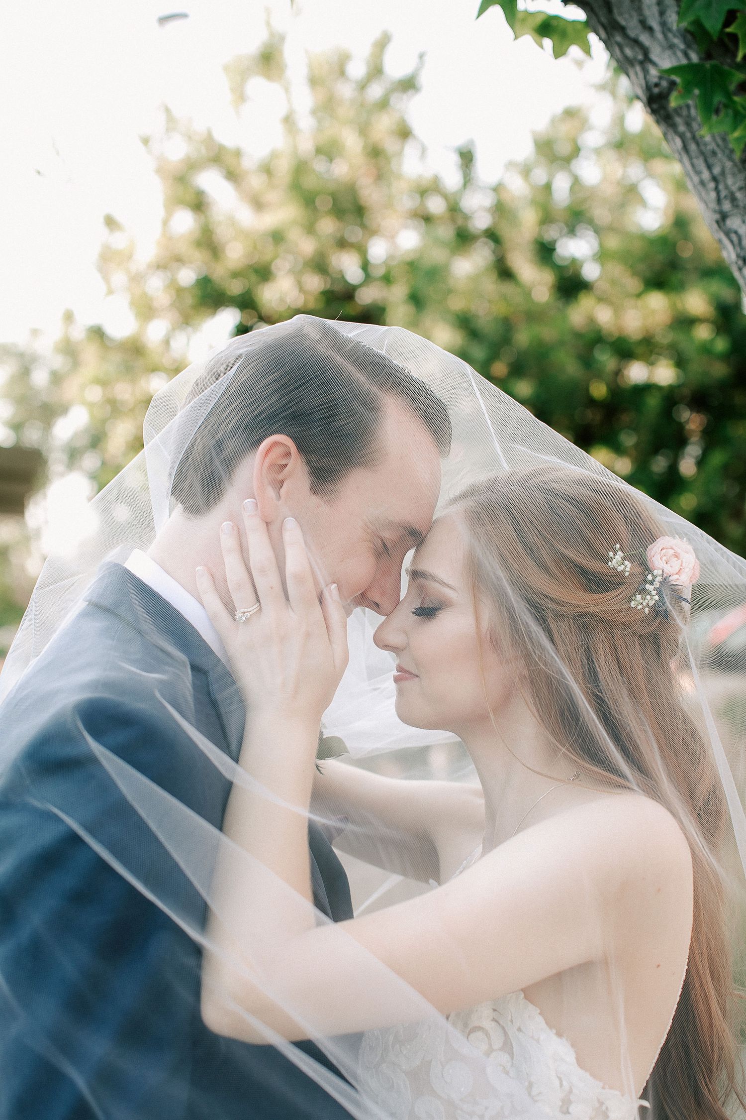 Bride and groom share an intimate moment under a wedding veil outdoors, foreheads touching in a romantic pose.