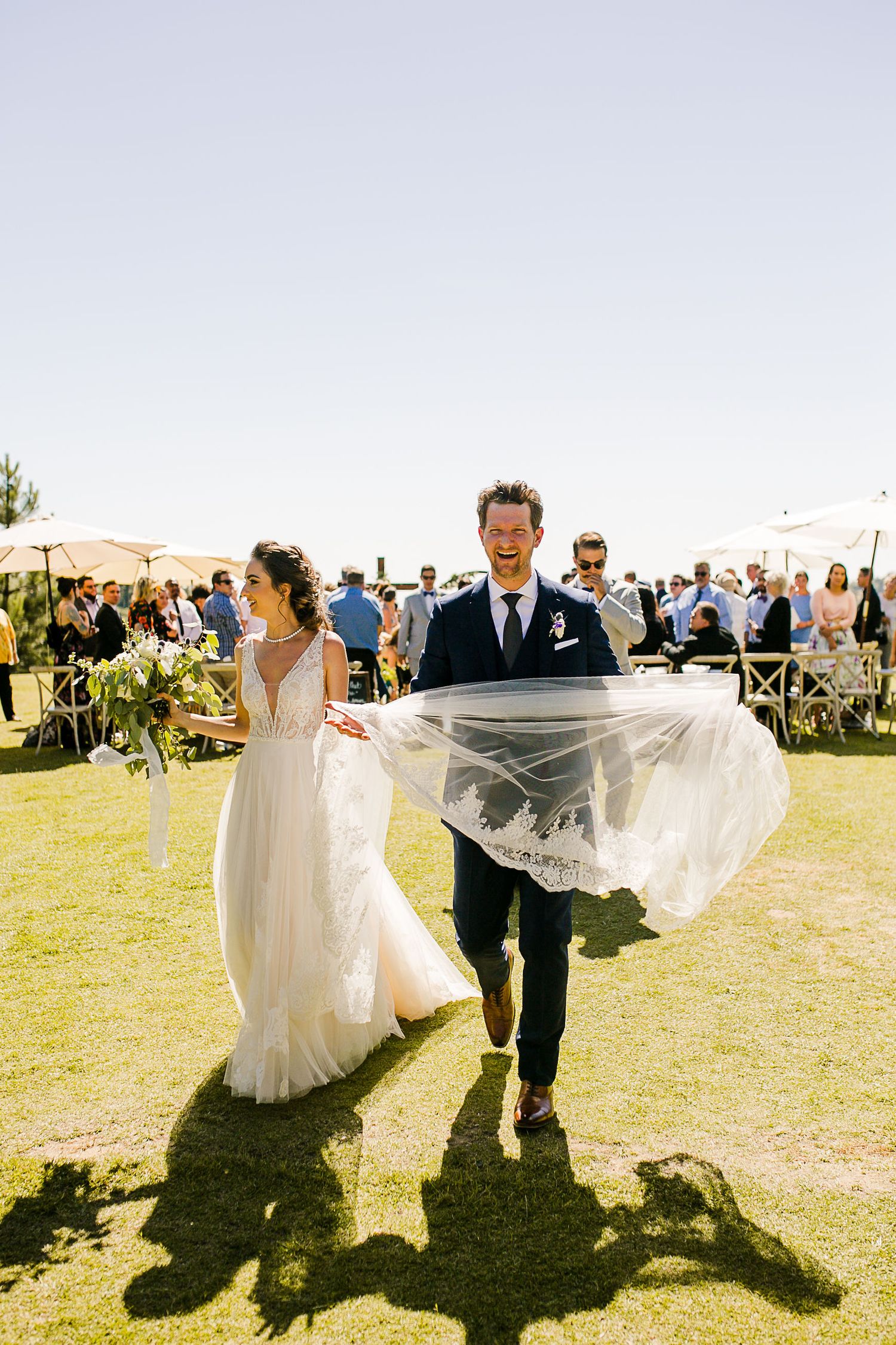 Joyful bride and groom walking down the aisle outdoors, veil flowing in the wind, guests seated behind them.