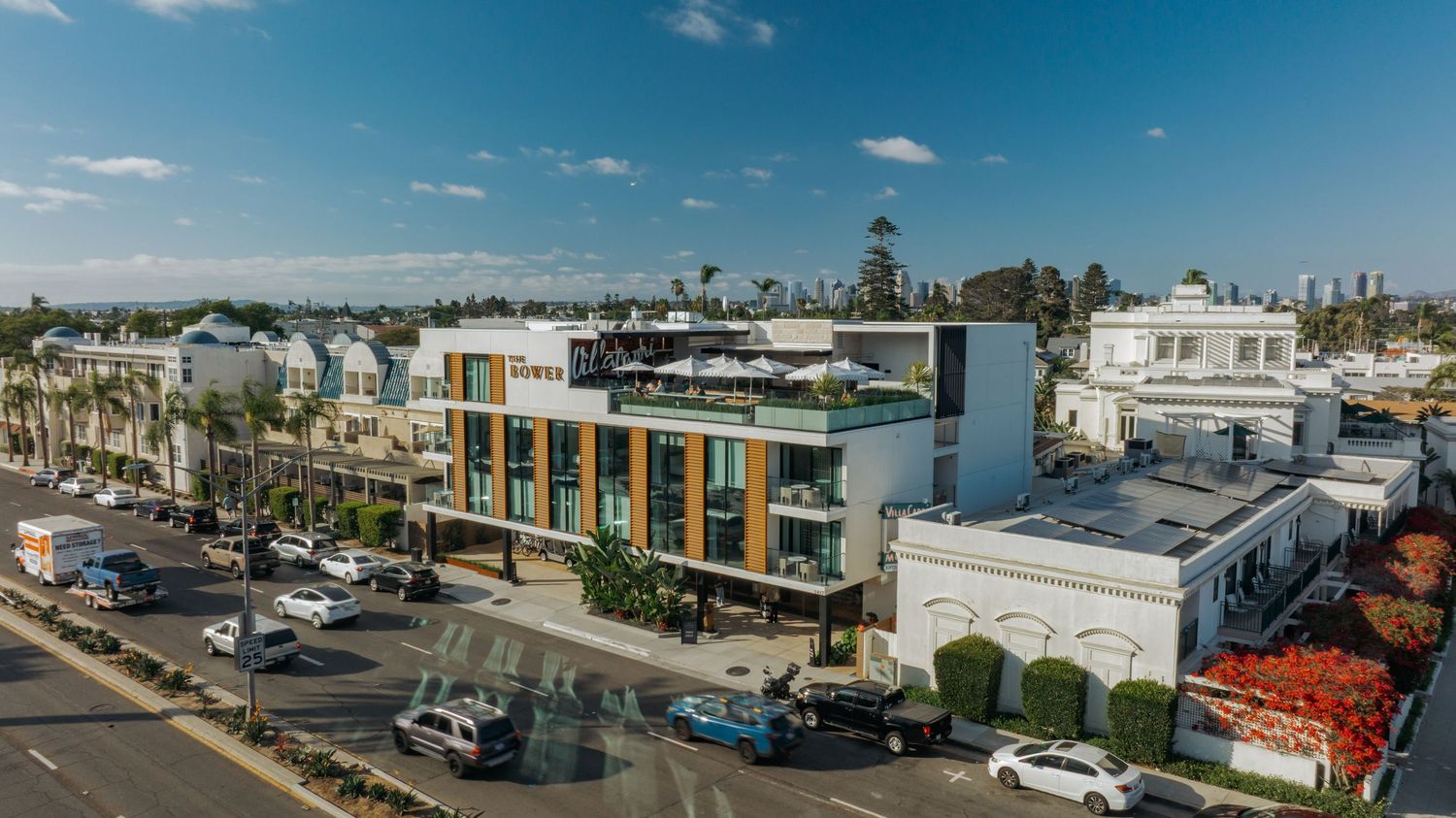 Aerial view of a modern mixed-use building on a busy coastal city street lined with palm trees and shops.