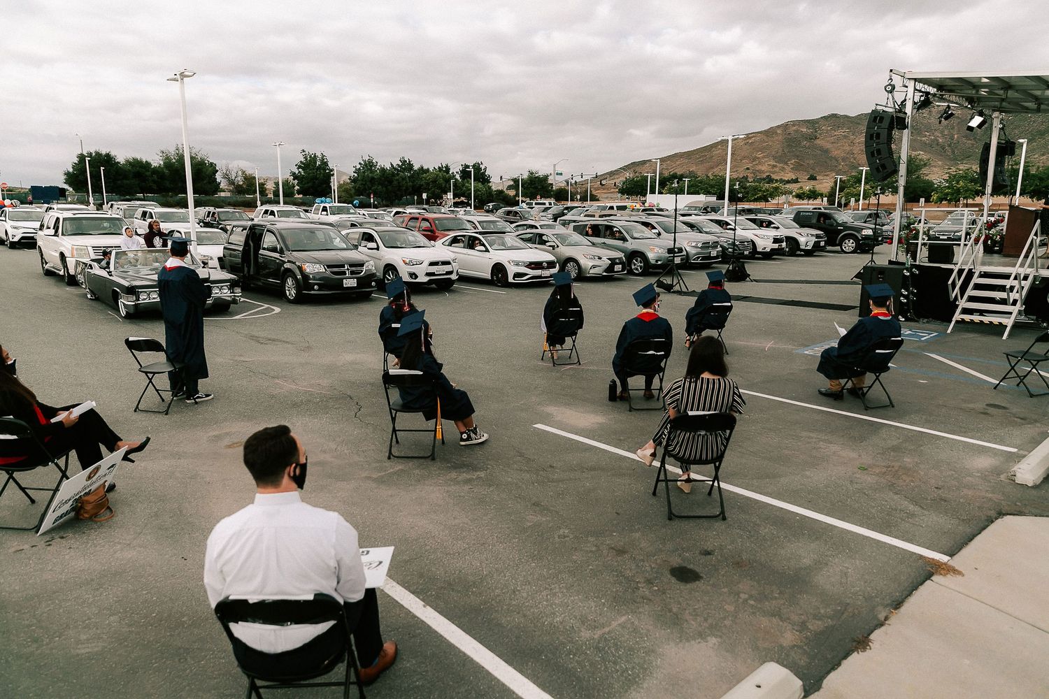 People sitting in chairs spaced apart in a parking lot during an outdoor socially distanced gathering with cars in the background.
