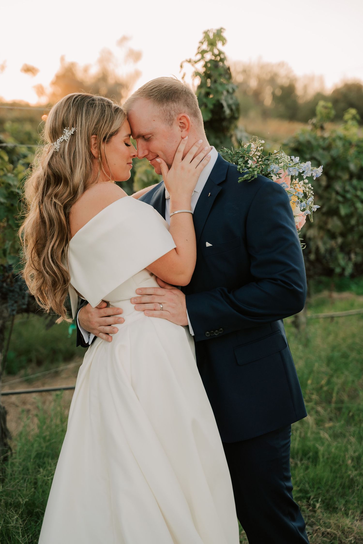 Bride and groom share romantic moment at golden hour in vineyard, touching foreheads in wedding attire.