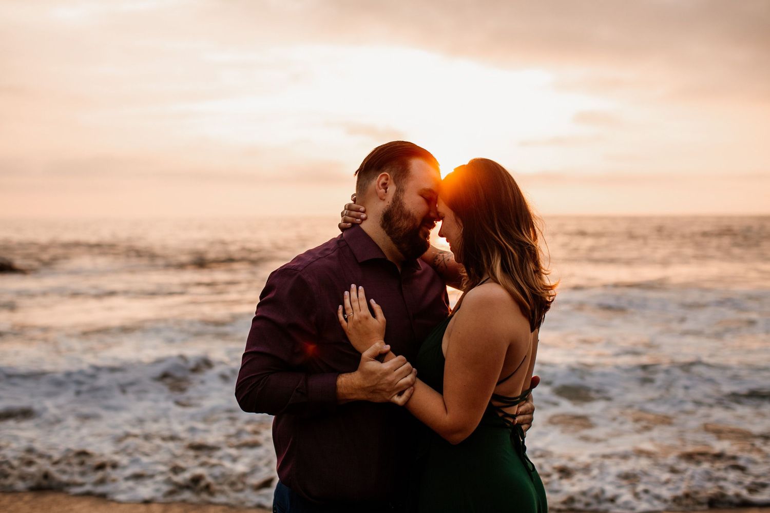 Couple sharing a romantic sunset moment by the ocean, man in burgundy shirt and woman in green dress.