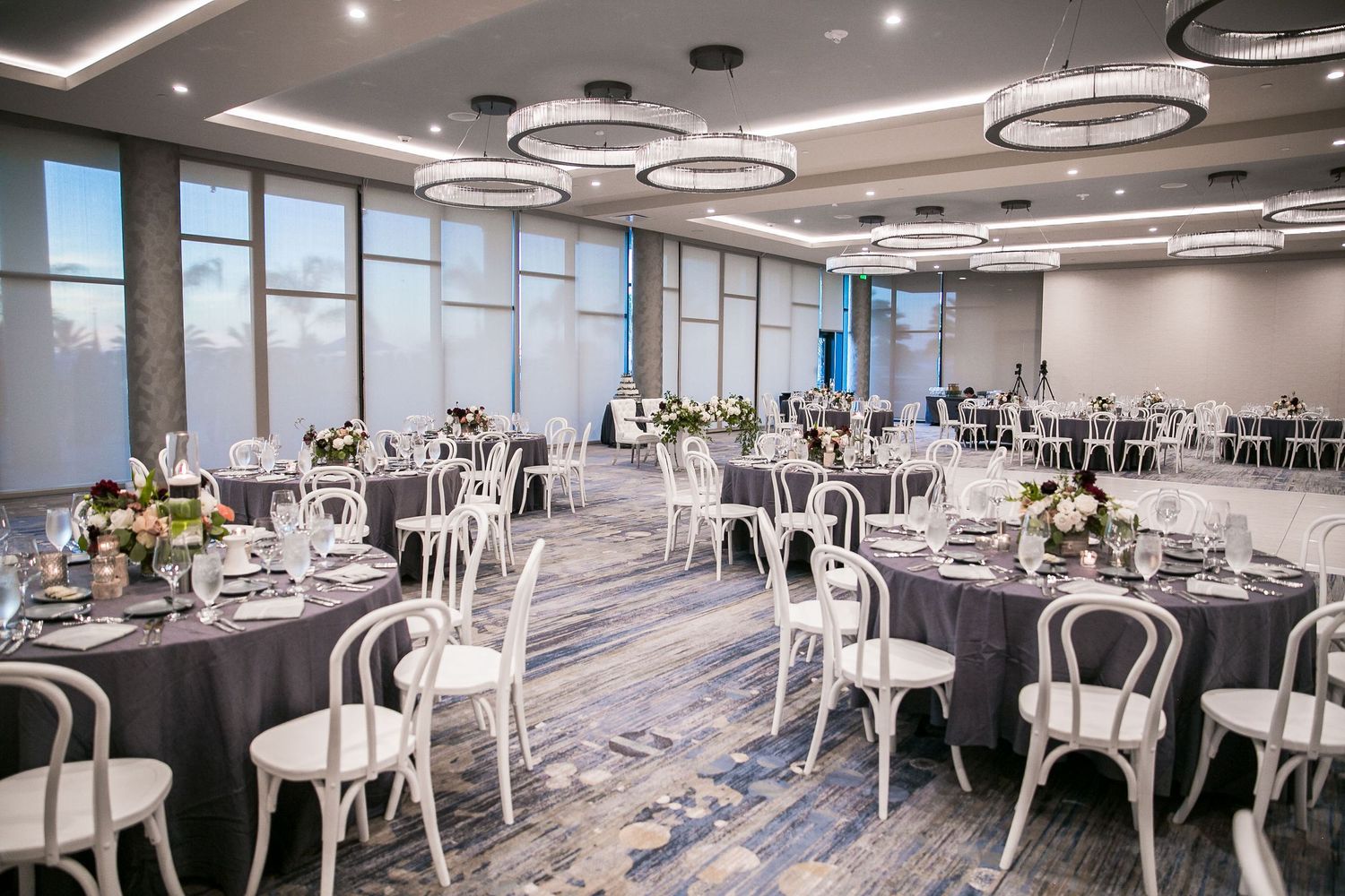 Elegant ballroom with round tables draped in gray linens, white chairs, floral centerpieces, and modern circular chandeliers.