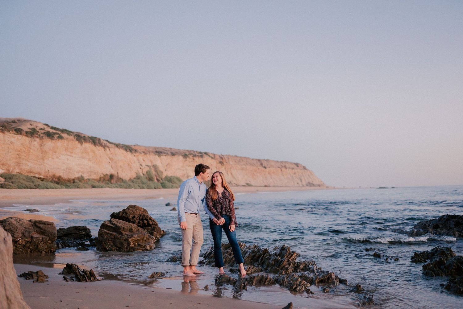 Couple standing on rocky beach shore at sunset with dramatic cliffs in background.