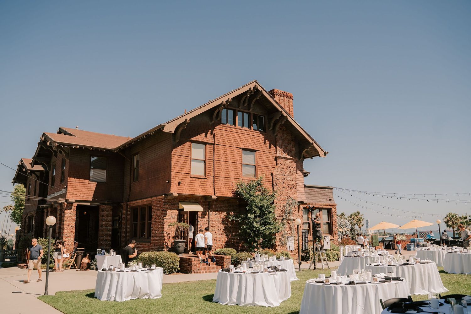 Outdoor wedding reception with white-clothed round tables on a green lawn beside a historic red brick craftsman venue under a blue sky.