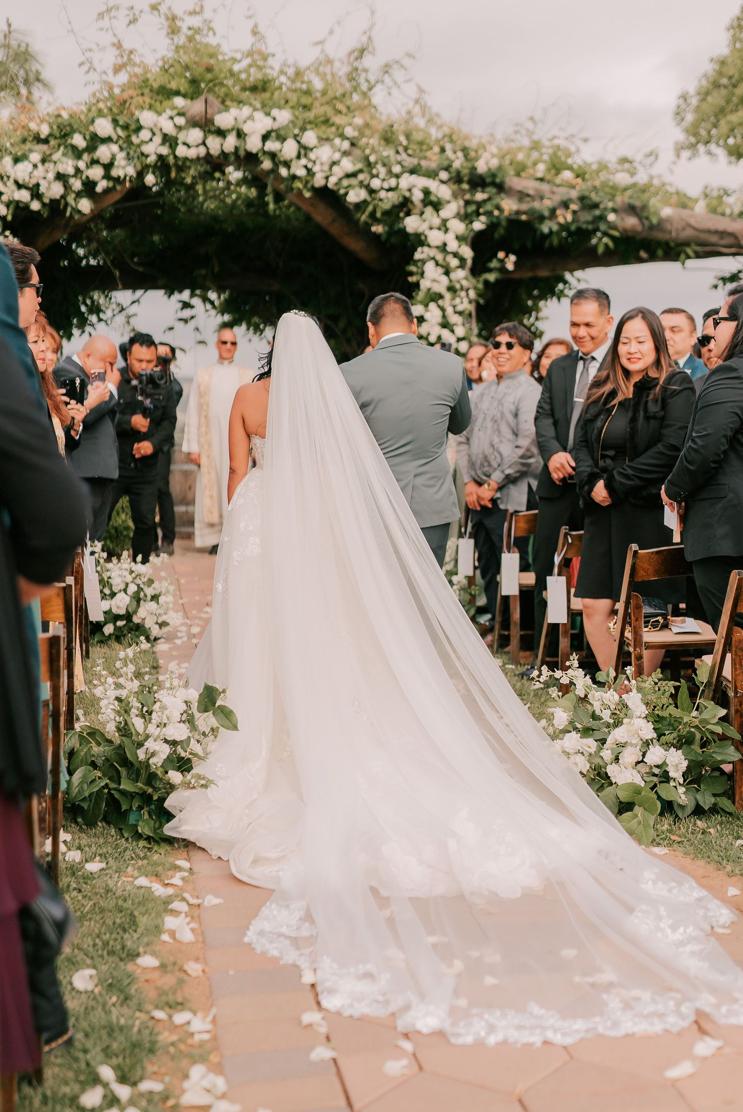 Bride walking down the aisle in a stunning white gown with a long lace-trimmed veil at an outdoor wedding ceremony.
