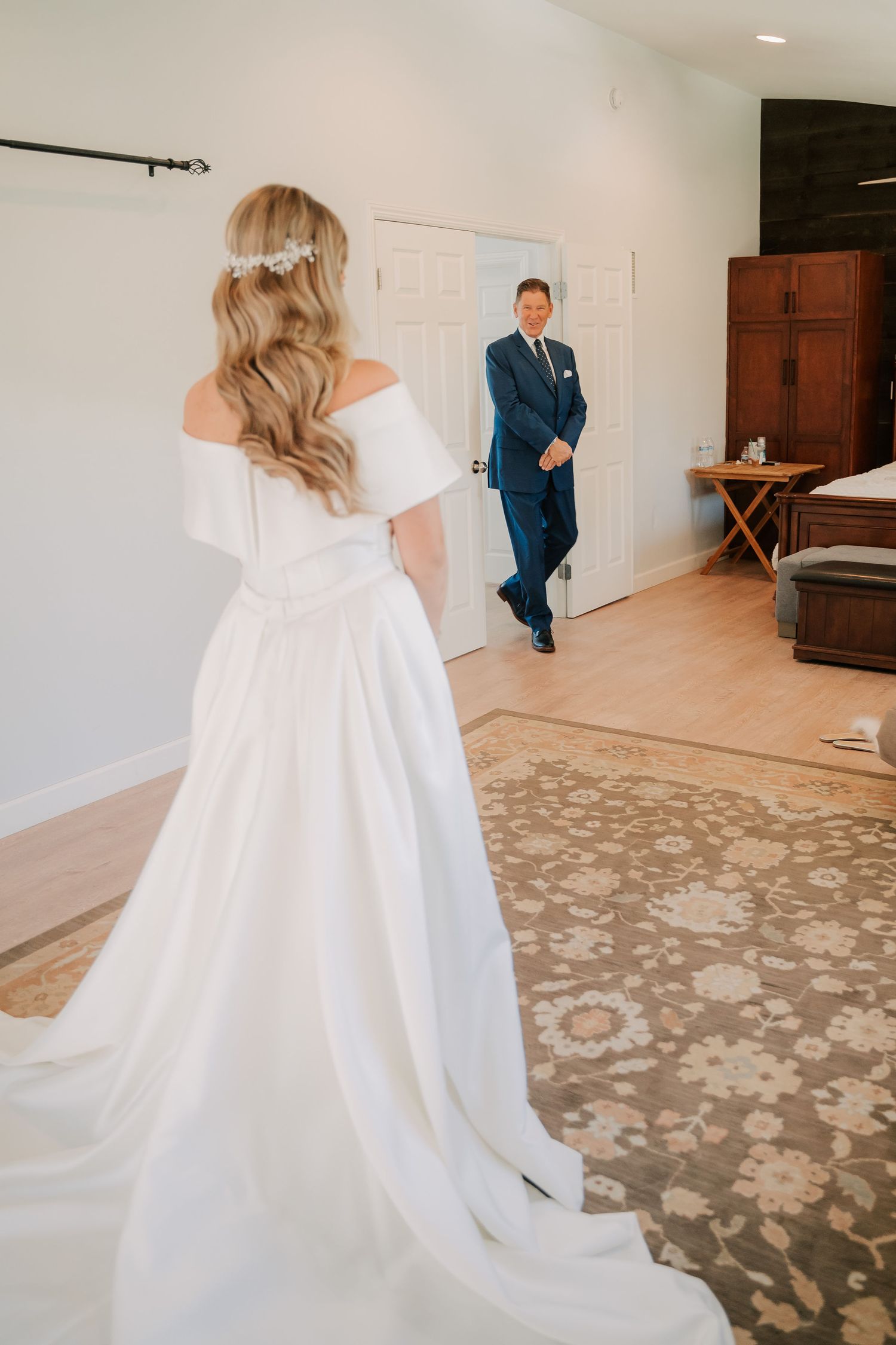 Bride in white off-shoulder gown with floral hair piece shares first look moment with groom in navy suit.