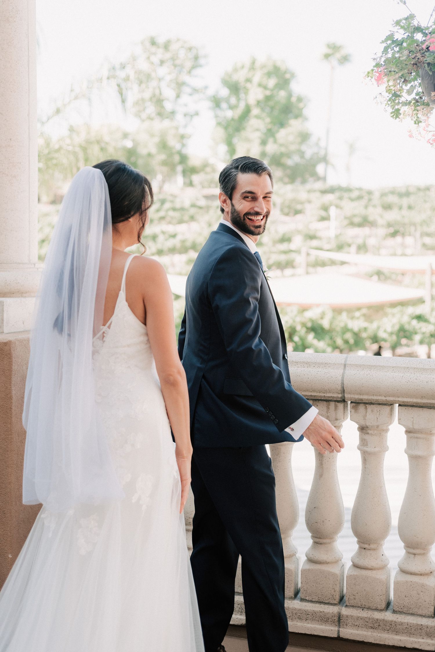 Smiling groom in navy suit holds bride's hand on elegant balcony, bride wearing white lace wedding gown and veil.