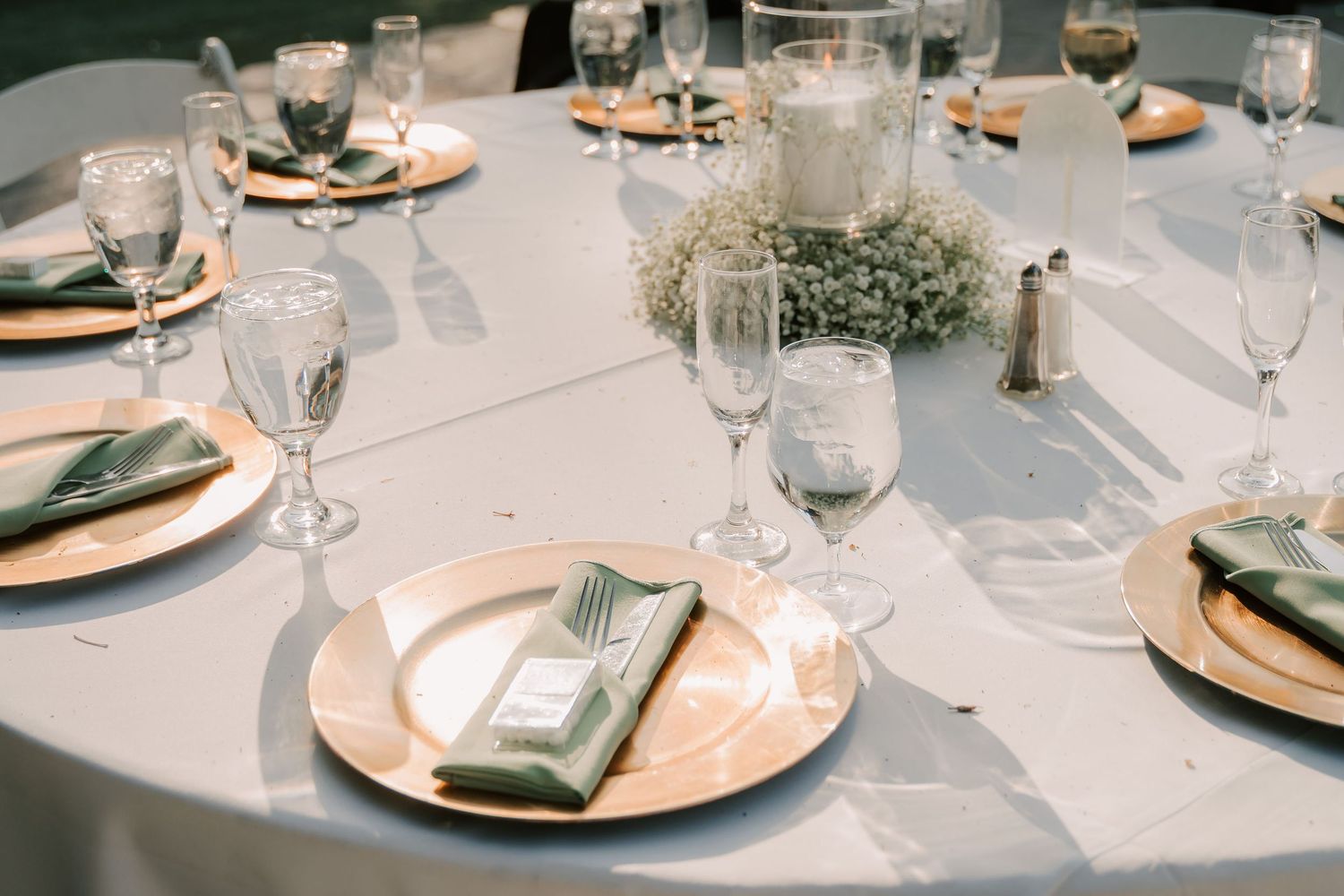 Elegant wedding reception table setting with gold charger plates, green napkins, crystal glasses, and baby's breath centerpiece.