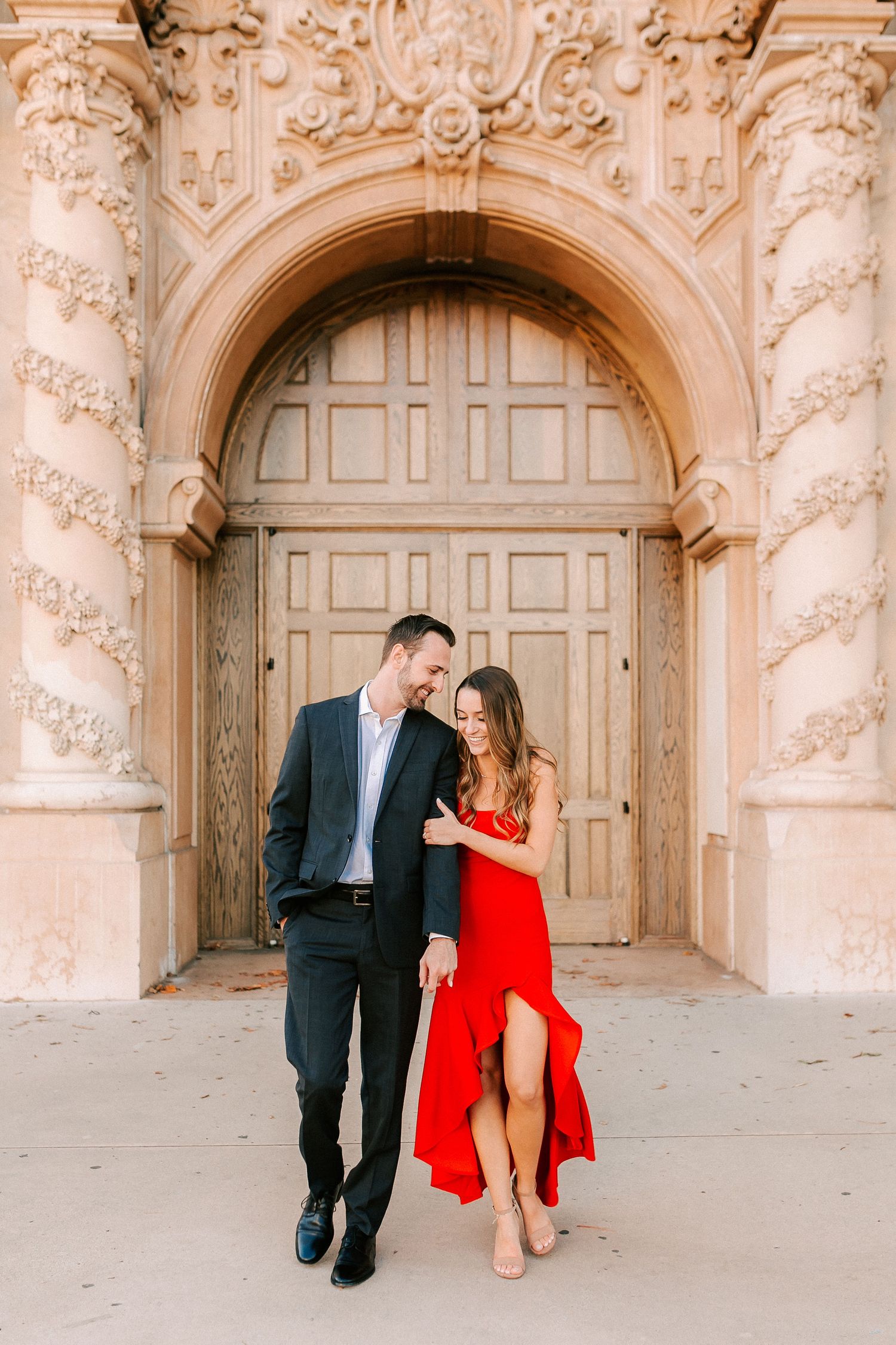 Couple posing in front of ornate arched doorway, woman in red dress and man in black suit.